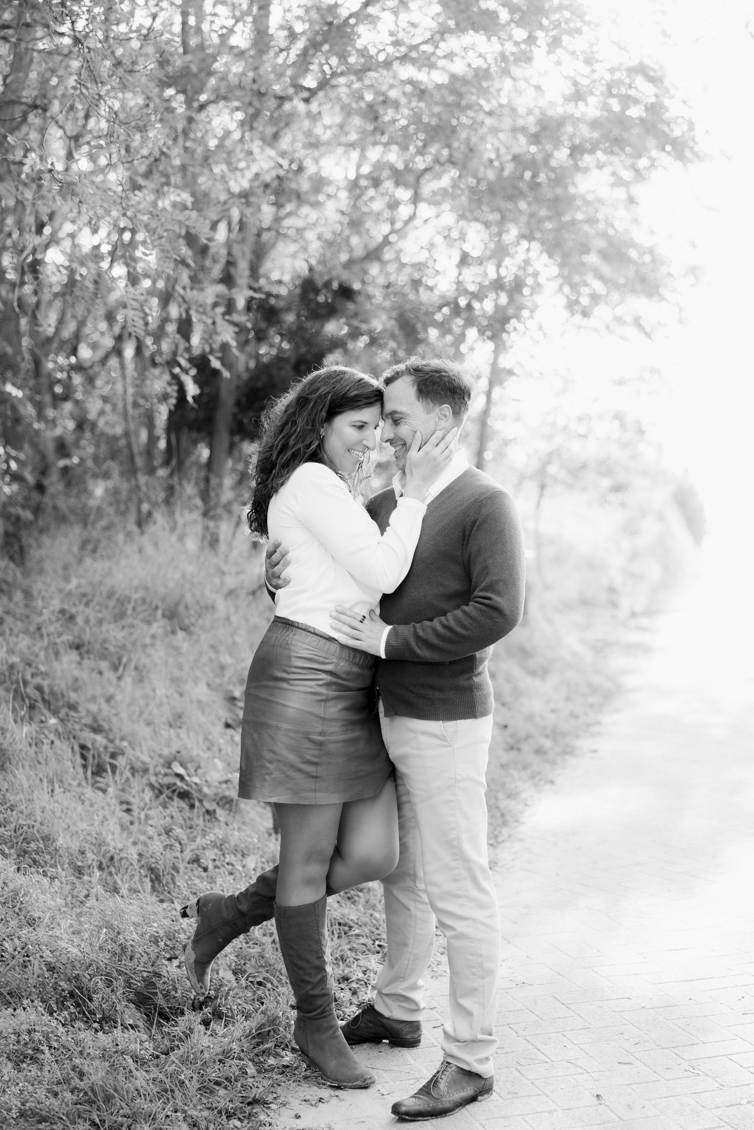 Black and white photo of a couple embracing and smiling on a path surrounded by trees.