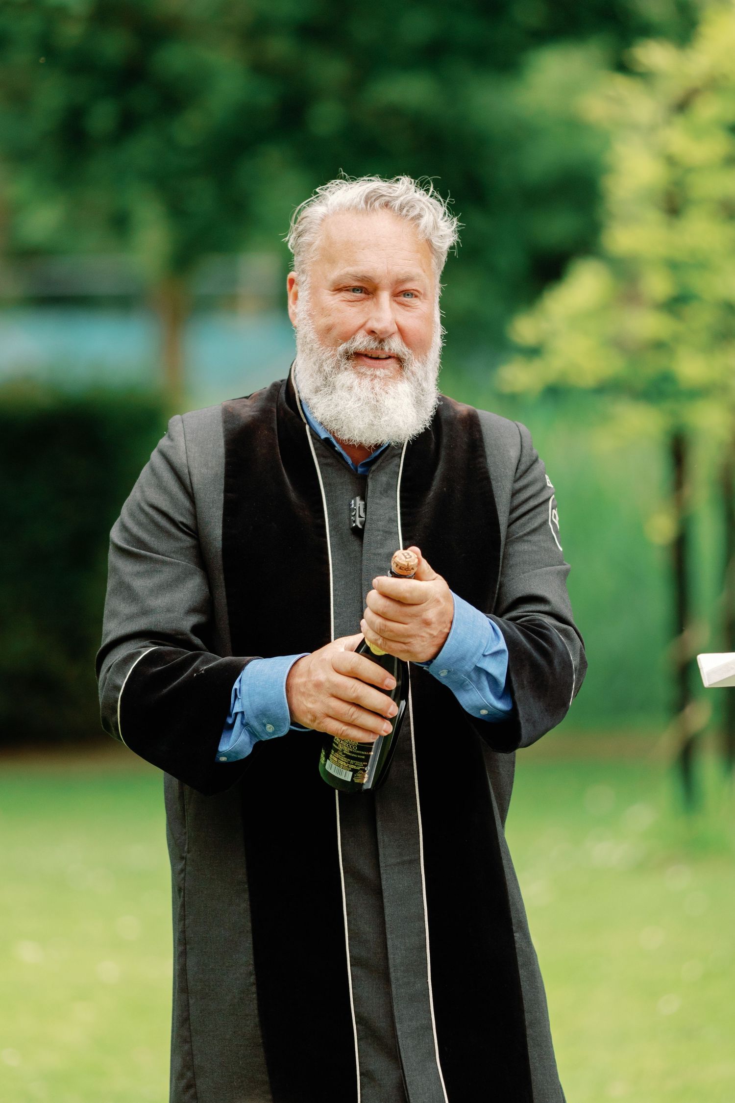 A person in black attire with a beard opens a bottle in an outdoor setting with trees in the background.