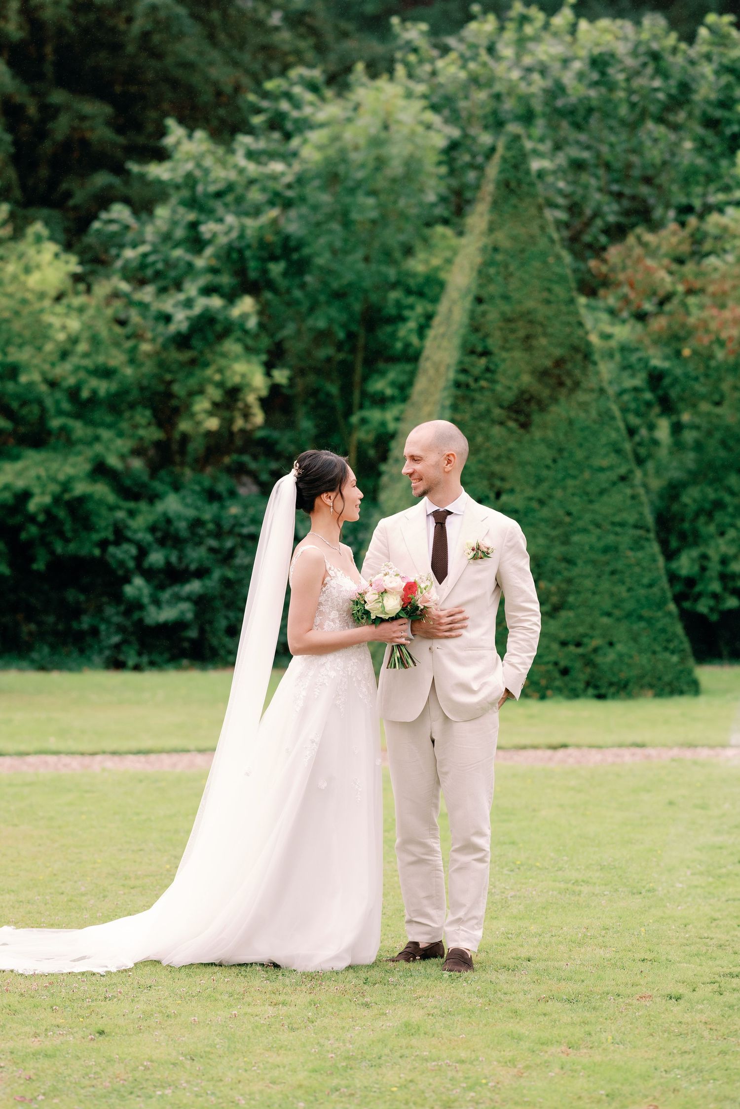 A bride and groom share an intimate moment on a lush green lawn with manicured topiary trees in the background.