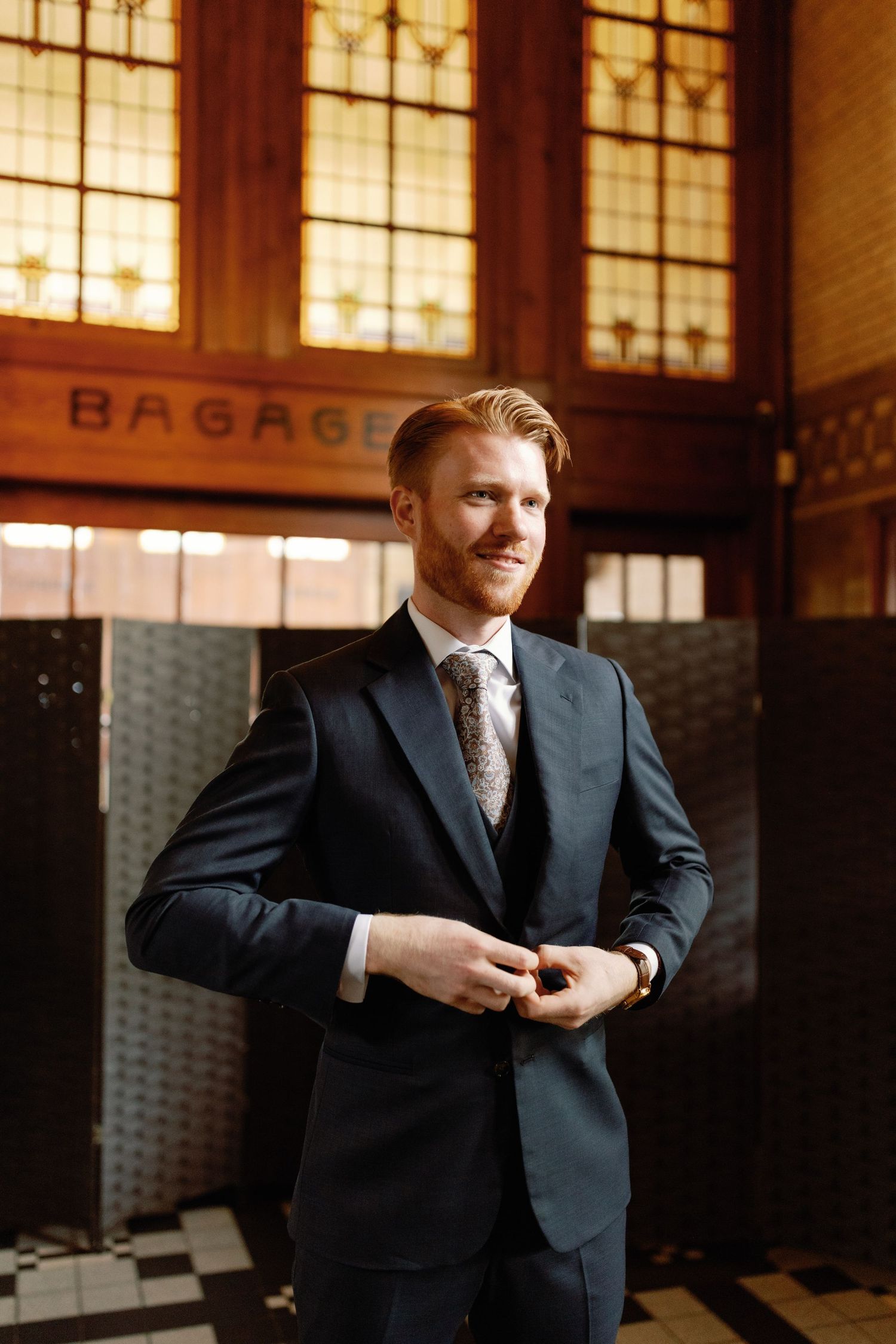 A person in a dark suit adjusts their tie while standing in a vintage train station with illuminated windows behind them.
