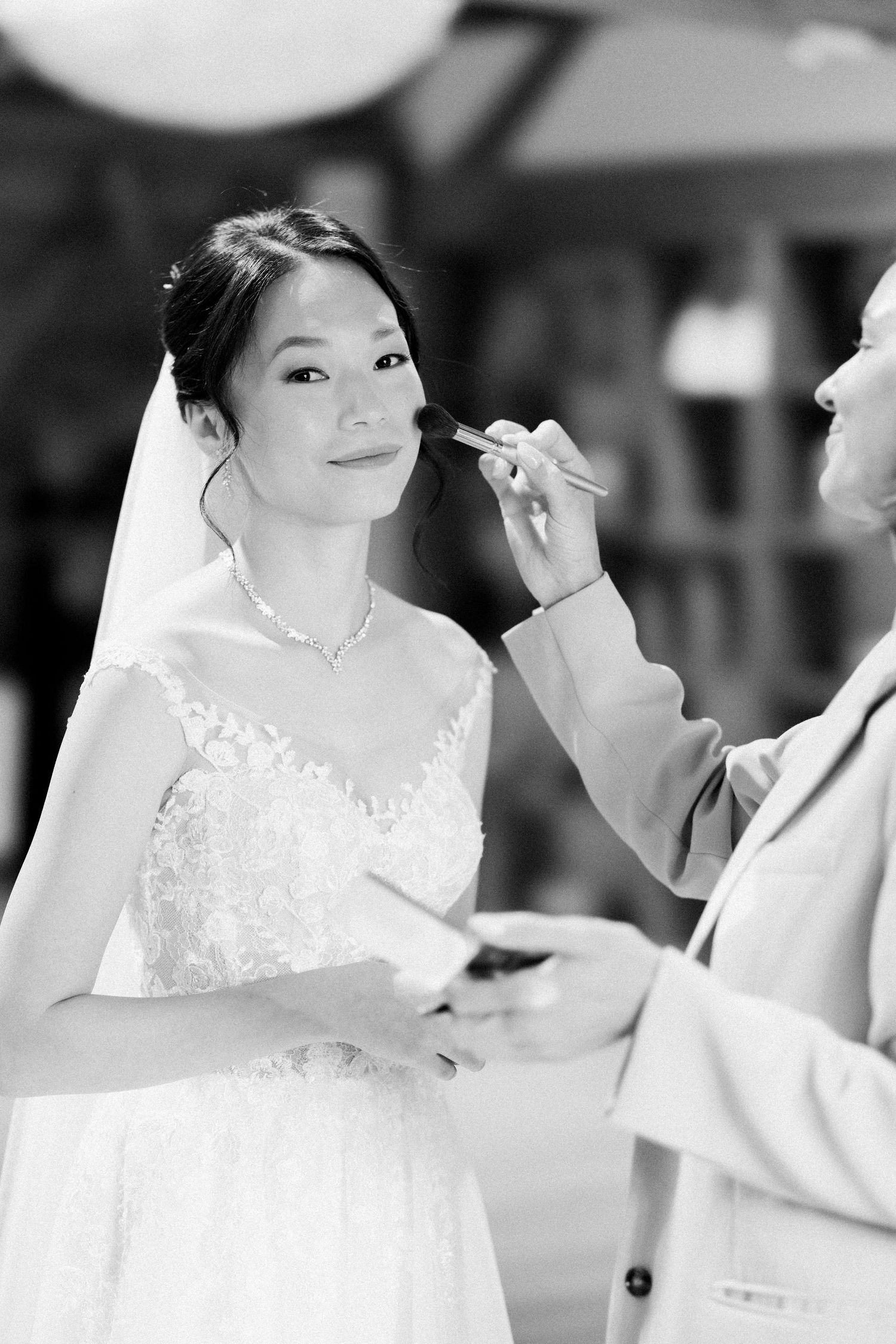 Black and white photo of a bride in a lace wedding dress getting makeup applied during wedding preparations.