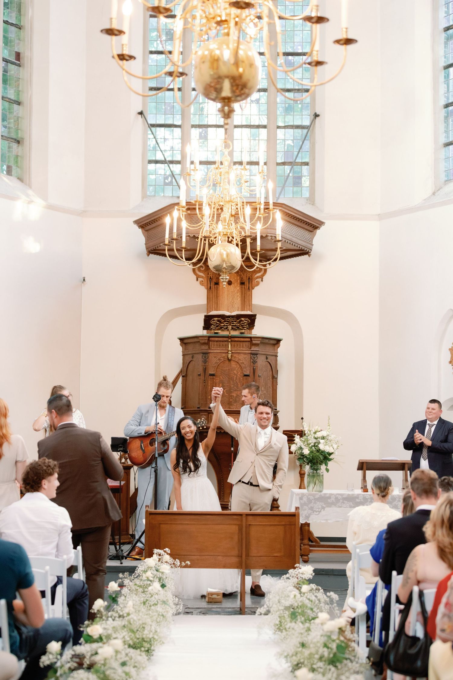 A bright church wedding ceremony with elegant chandeliers, white floral arrangements and guests seated in wooden pews.