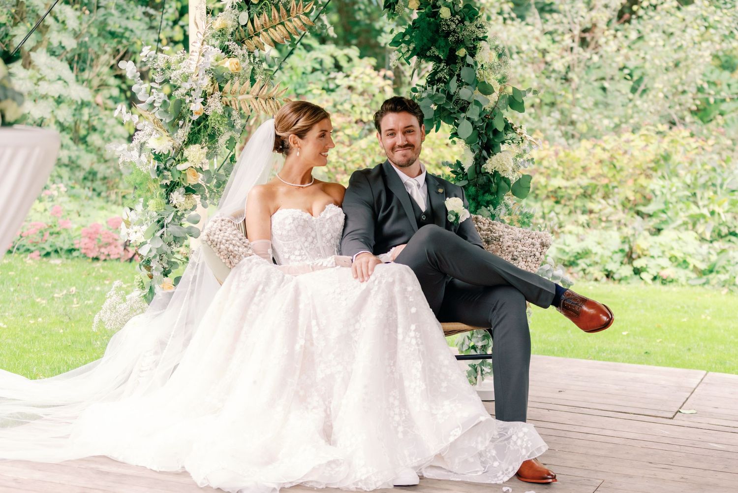 A couple in formal wedding attire sits together on wooden steps in a lush garden setting.