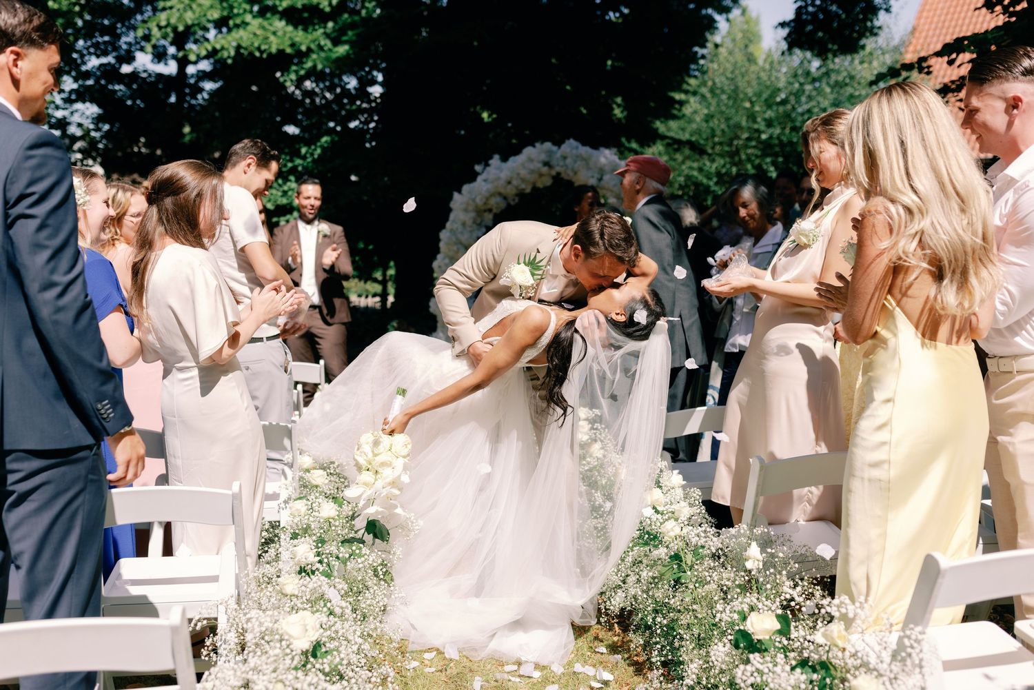 Wedding guests in pastel attire gather outdoors during a celebration with floral arrangements along the aisle.
