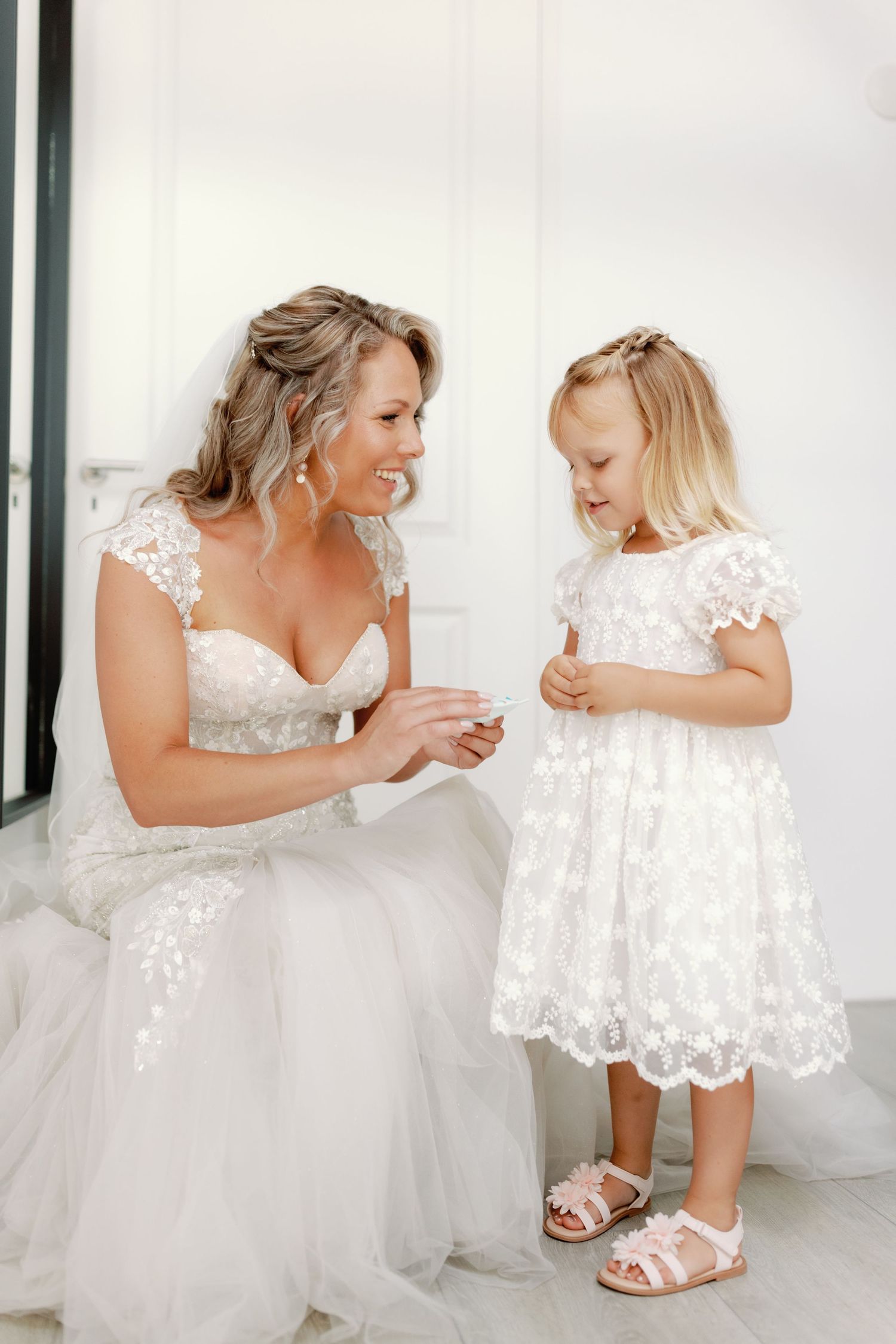 Smiling bride in a lace wedding gown and veil kneels to share a sweet moment with a young flower girl in a white embroidered dress.