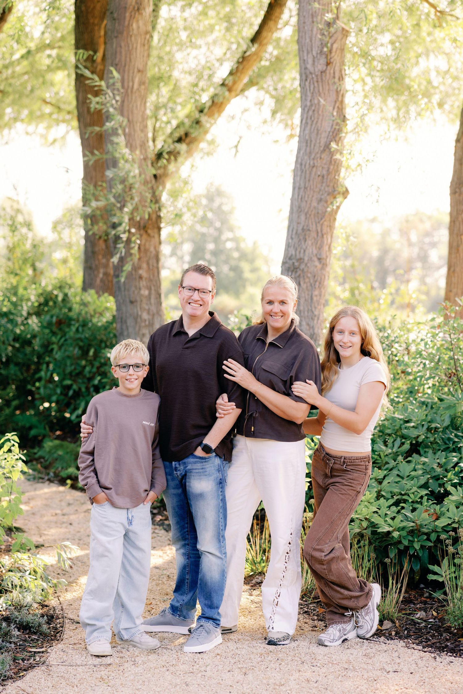 Family of four posing together on a garden path surrounded by tall trees and greenery, wearing casual outfits on a sunny day.