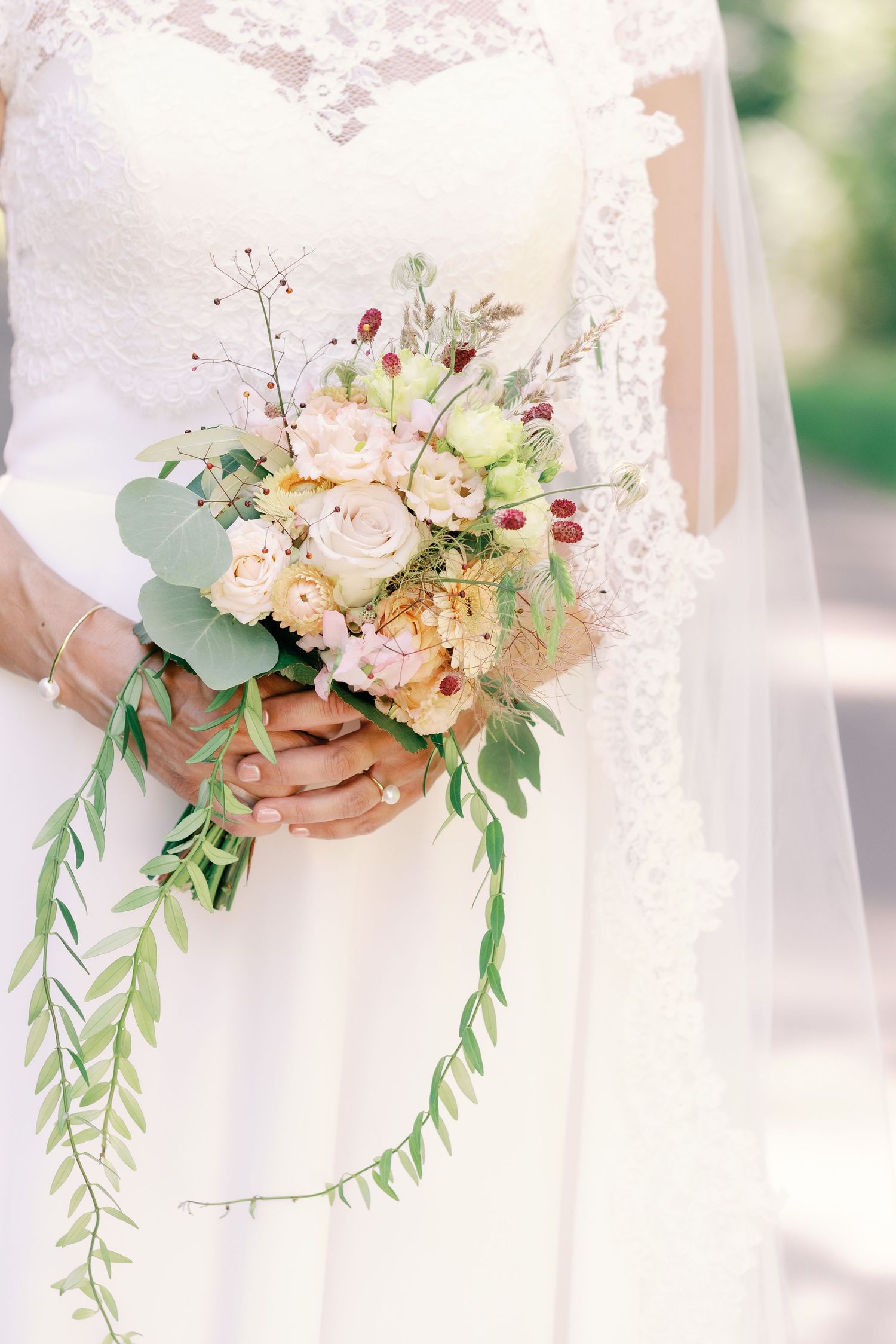 Delicate bridal bouquet with blush roses, greenery and berries held against a white lace dress background.