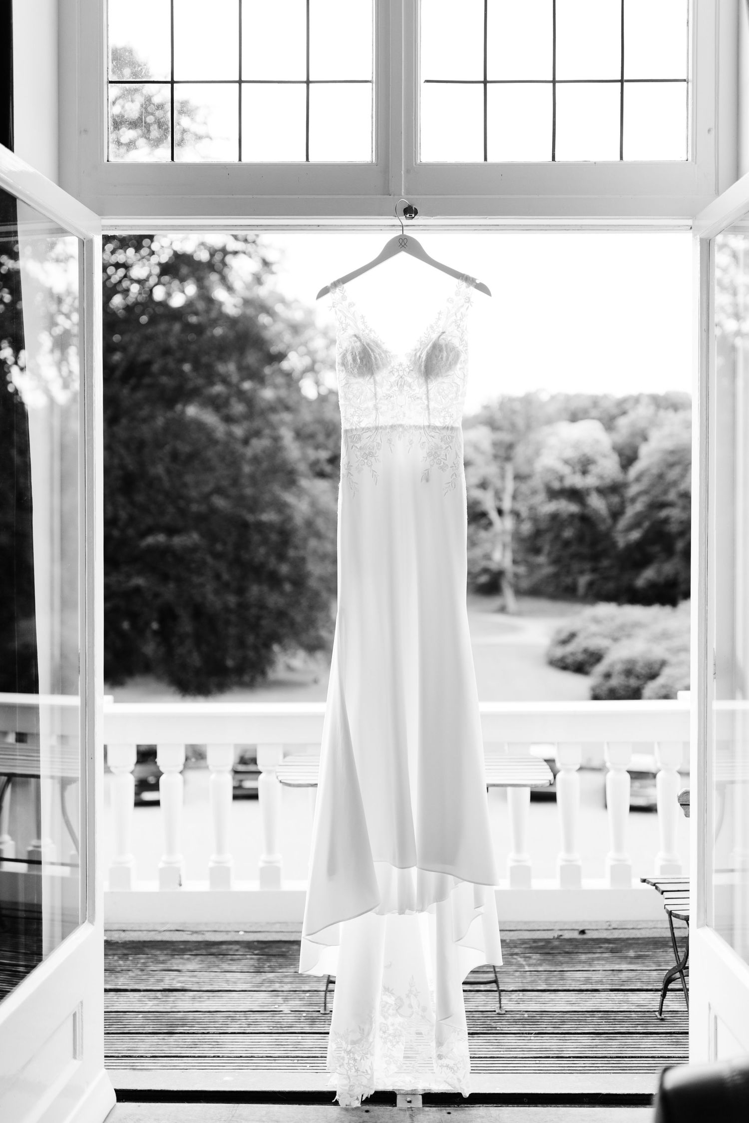An elegant white wedding dress hangs in a window frame overlooking a scenic outdoor porch with white railings.