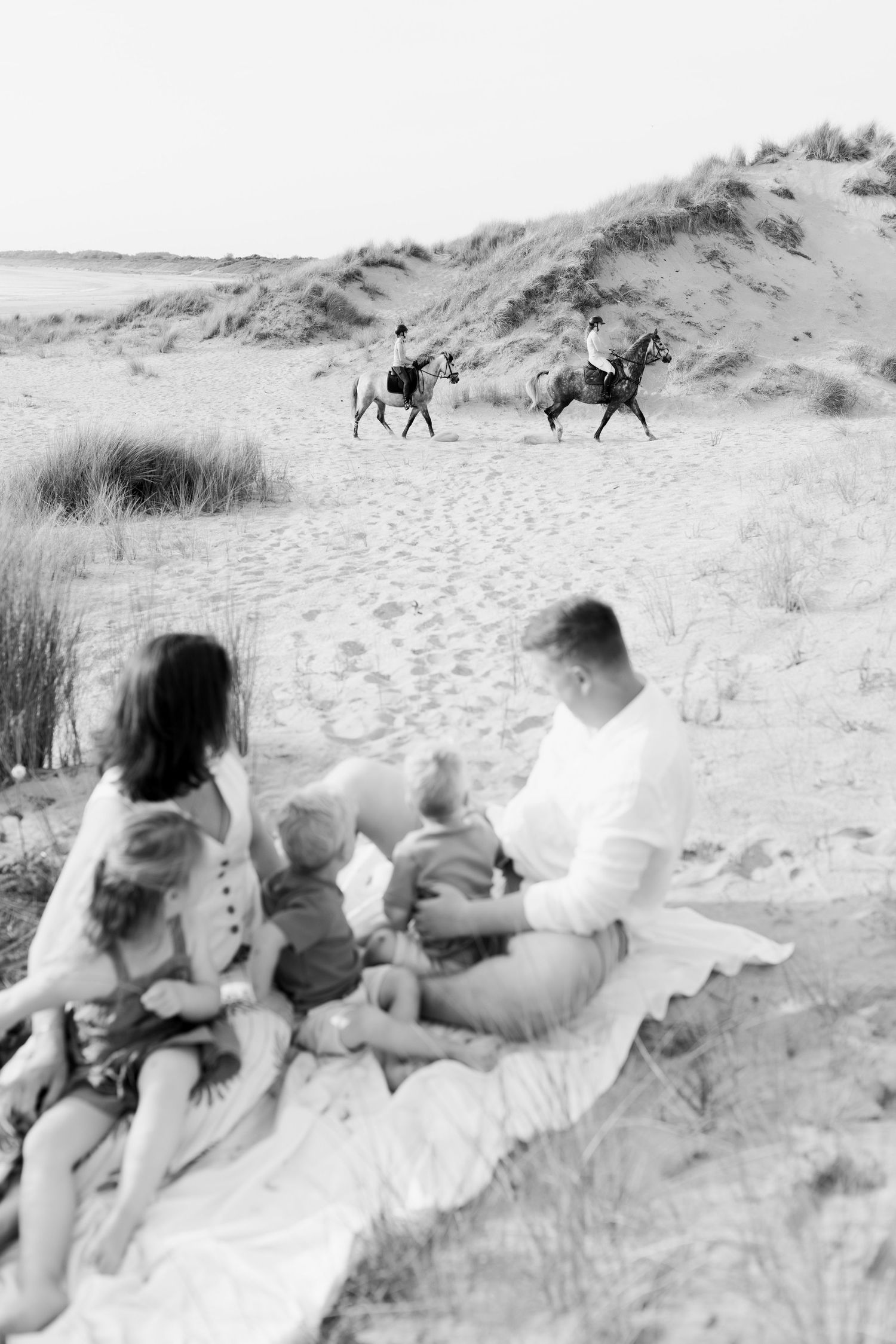 Black and white photo of a family sitting on a blanket on sandy beach dunes watching two horseback riders pass by.