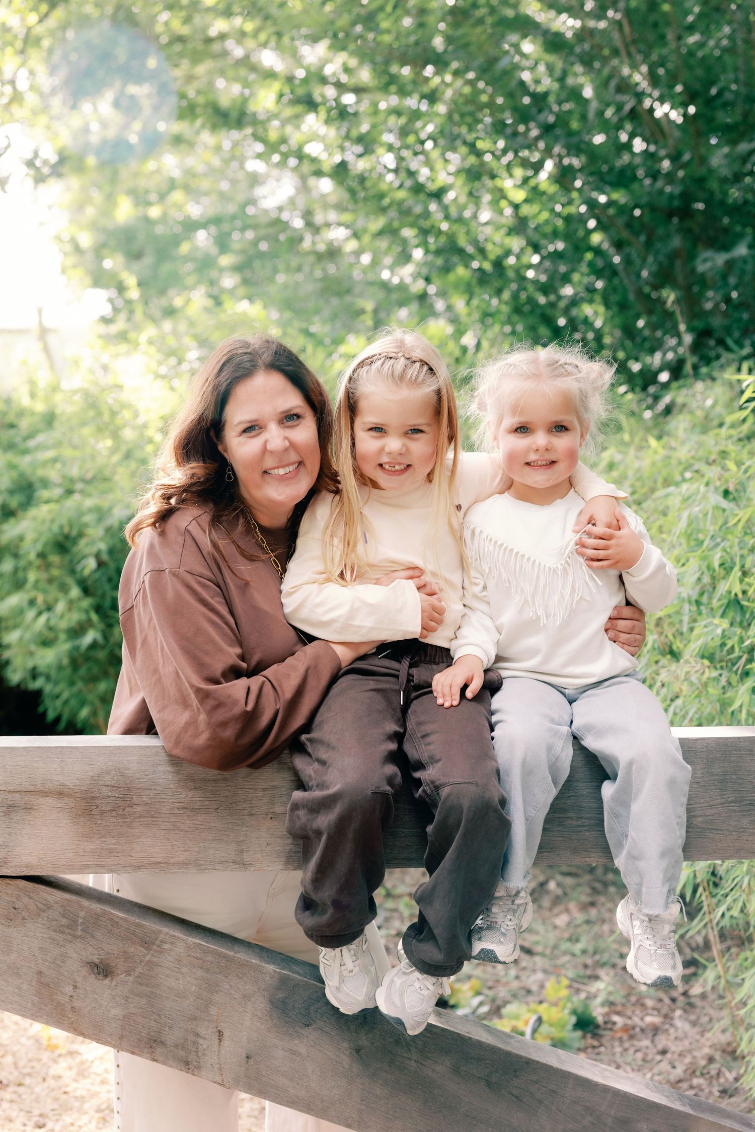 A smiling woman in a brown top sits on a wooden fence with two young girls in cream sweaters, surrounded by green trees.