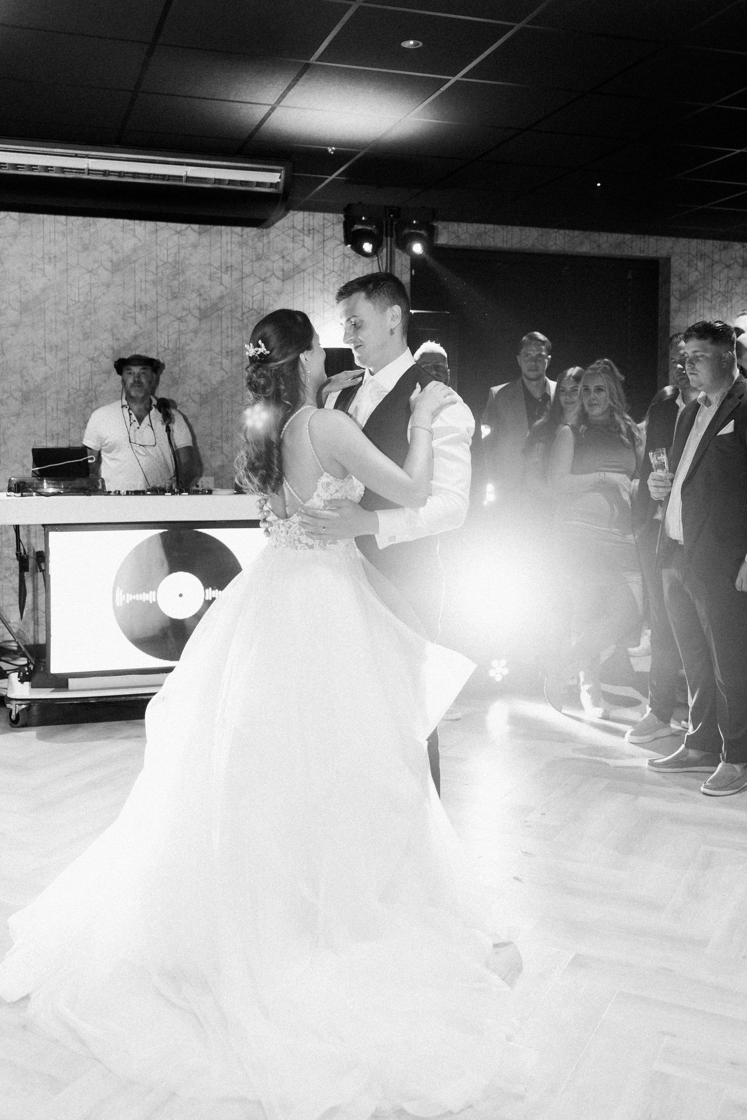 A couple shares their first dance at a wedding reception with a DJ booth visible in the background.