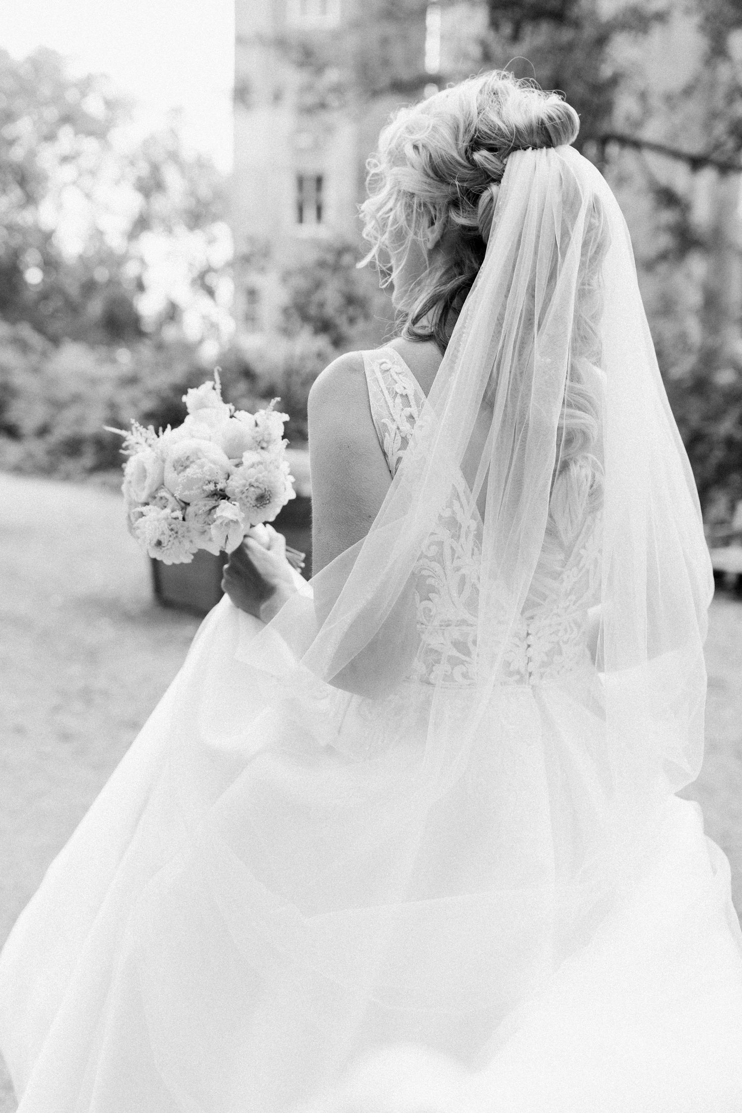A bride in a flowing white wedding gown and veil stands gracefully in a garden setting captured in black and white.