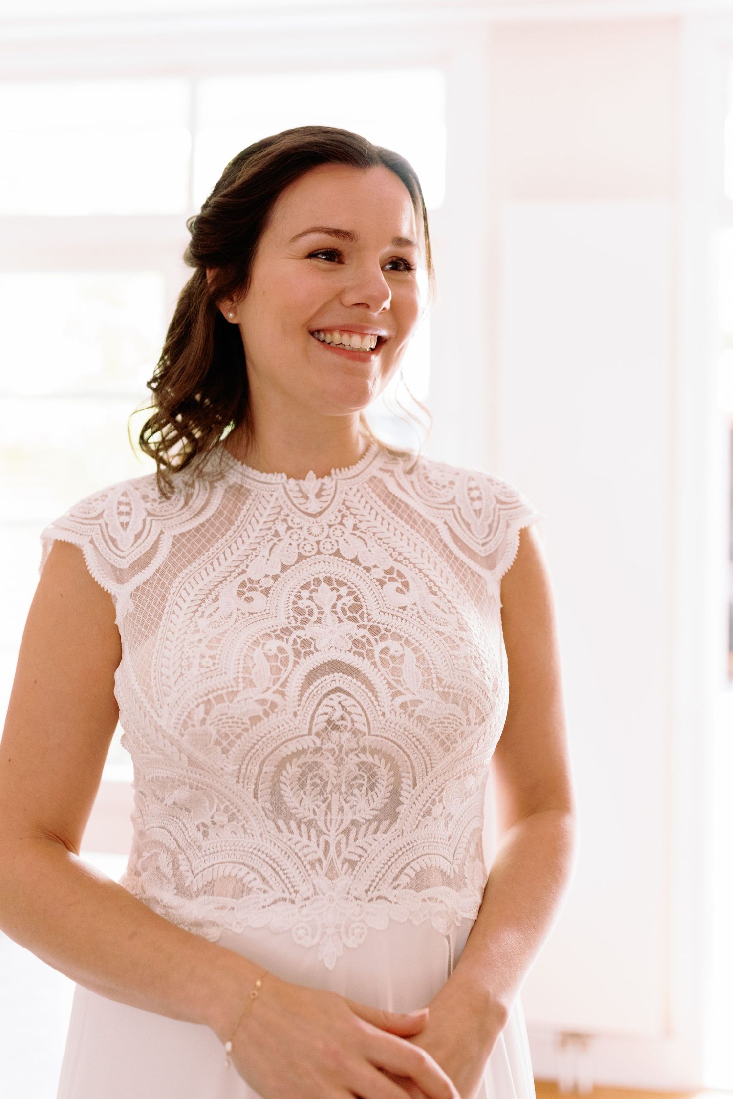 A person wearing an elegant white lace dress with a high neckline and delicate damask pattern smiles warmly.