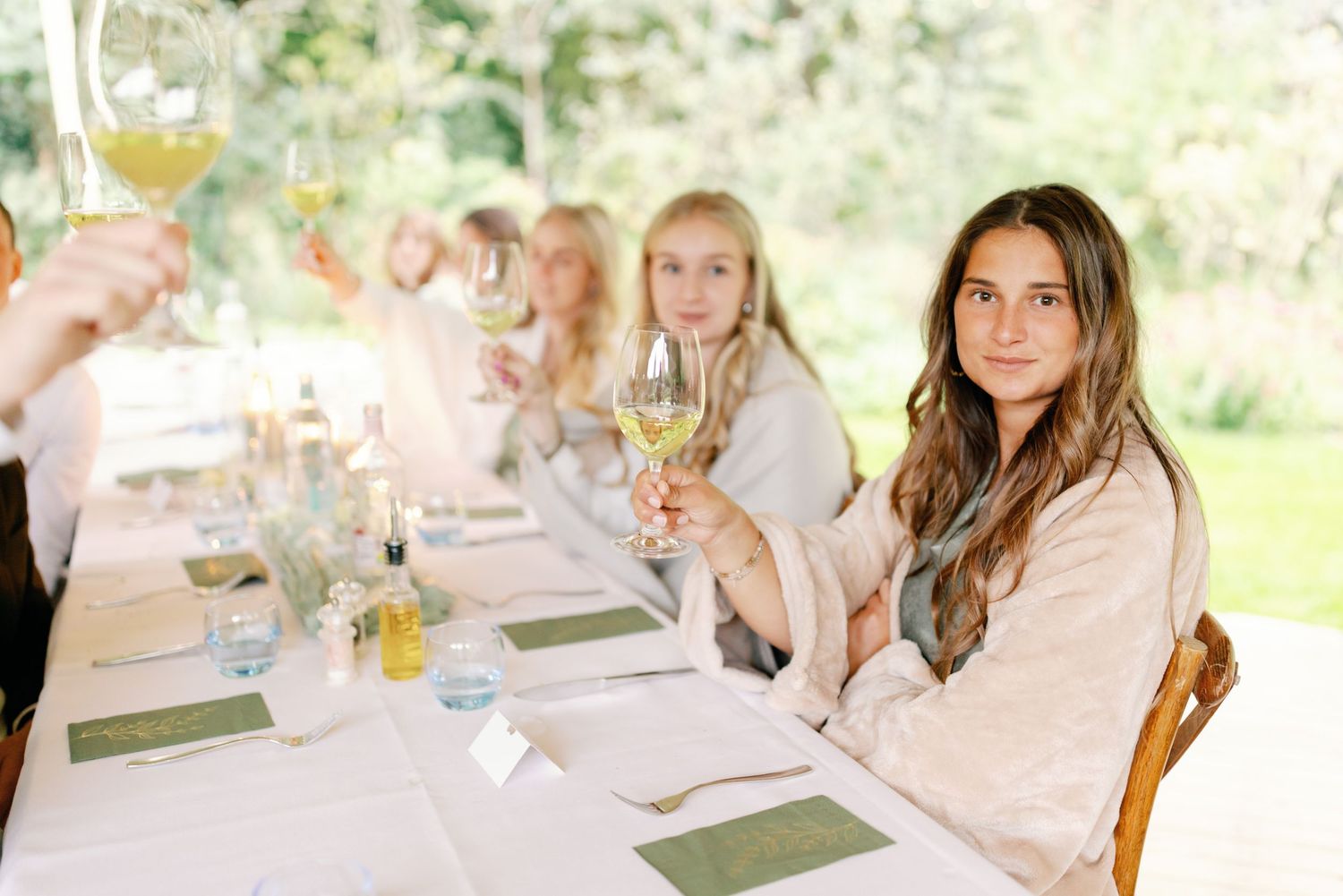 A group enjoys a casual outdoor dining experience at a long table set with wine glasses and plates in a sunny garden setting.