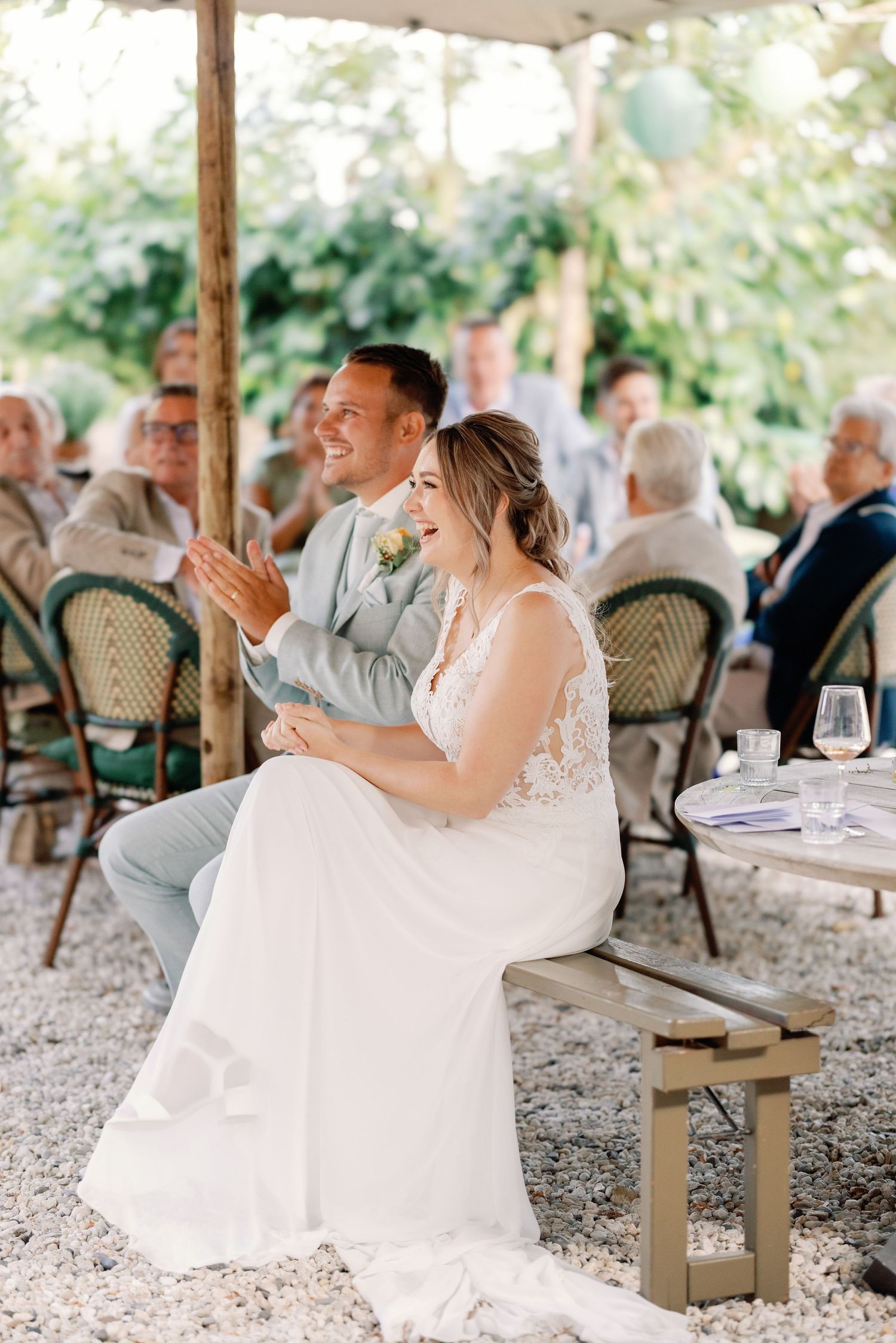 A couple in wedding attire laughs together while seated at an outdoor reception with guests in the background.