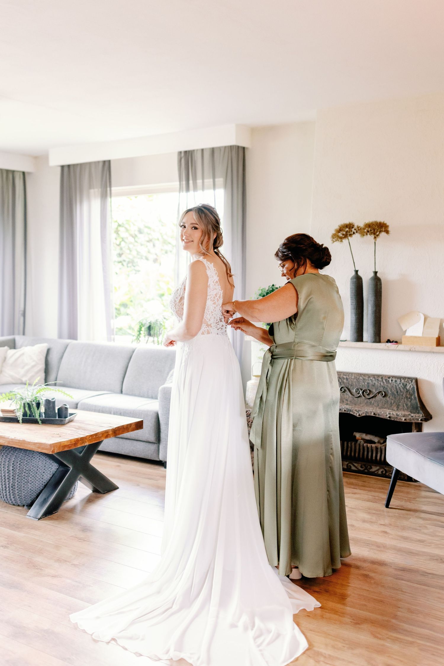 A bride in a flowing white wedding gown gets help with final adjustments from someone wearing a sage green dress in a modern room.