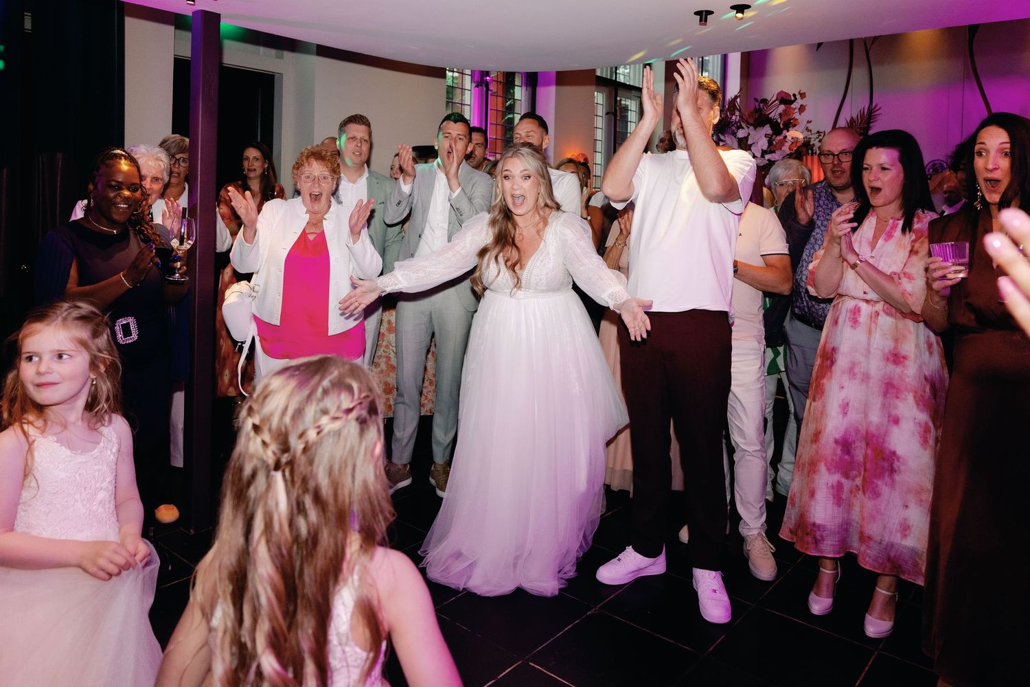 Wedding reception guests dance and celebrate on a dance floor illuminated with pink and purple lighting.