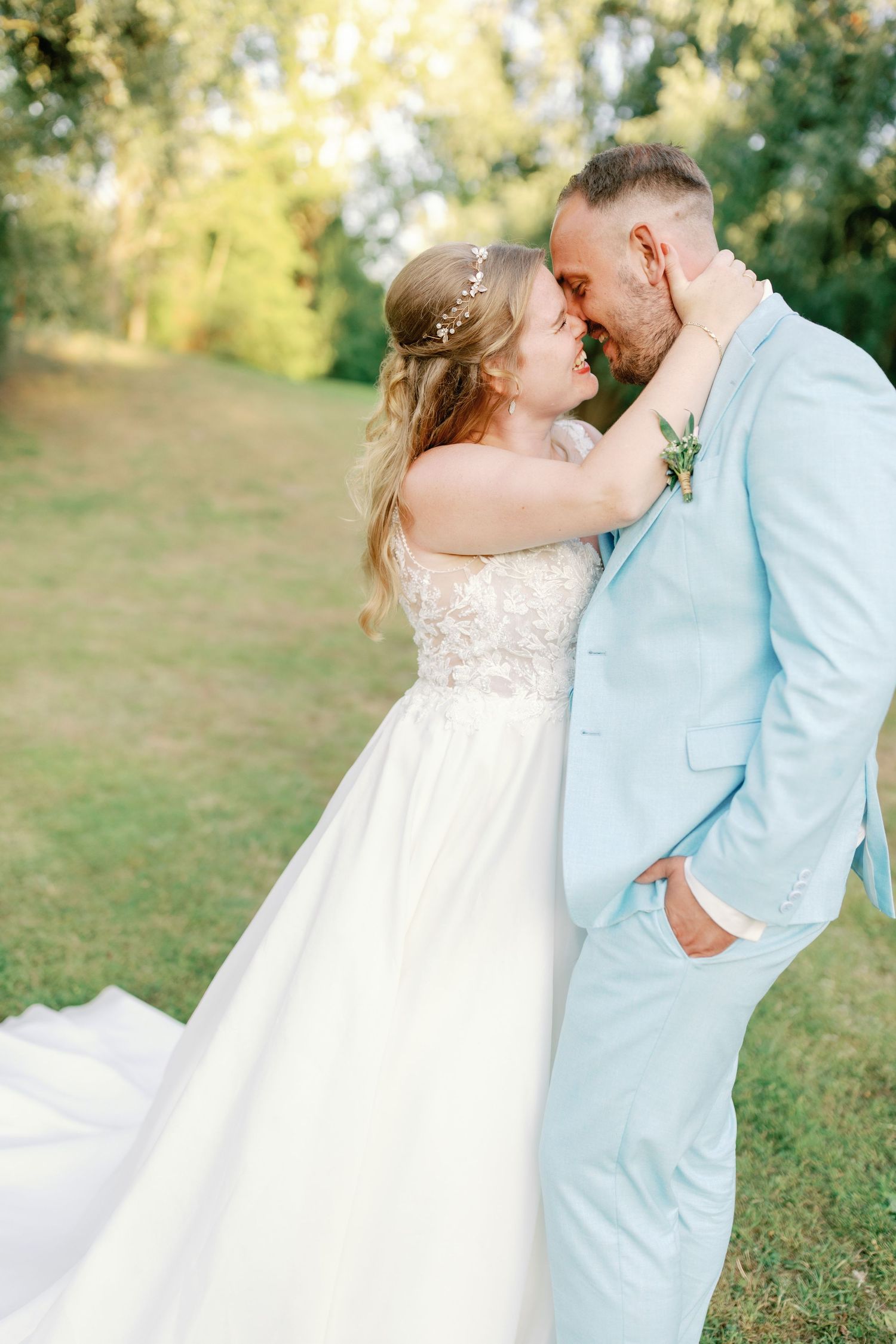 A couple in light blue and white wedding attire embrace romantically in a sunlit outdoor garden setting.