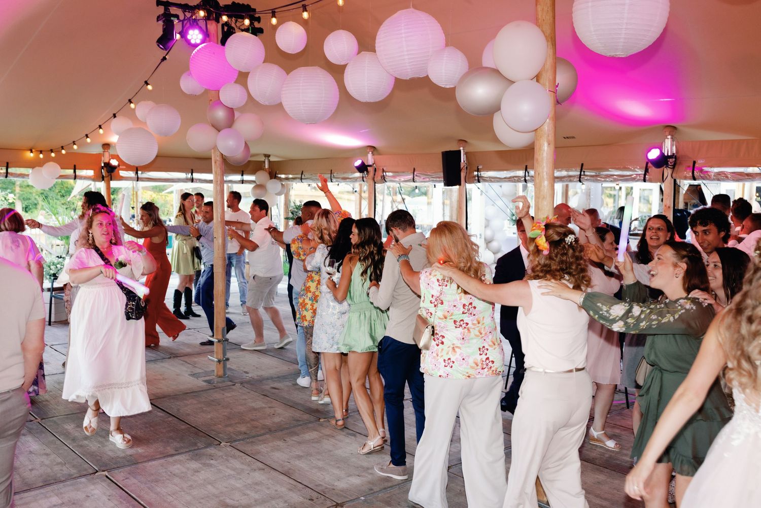 Wedding guests dancing and celebrating under a white tent with pink balloon decorations and lighting.