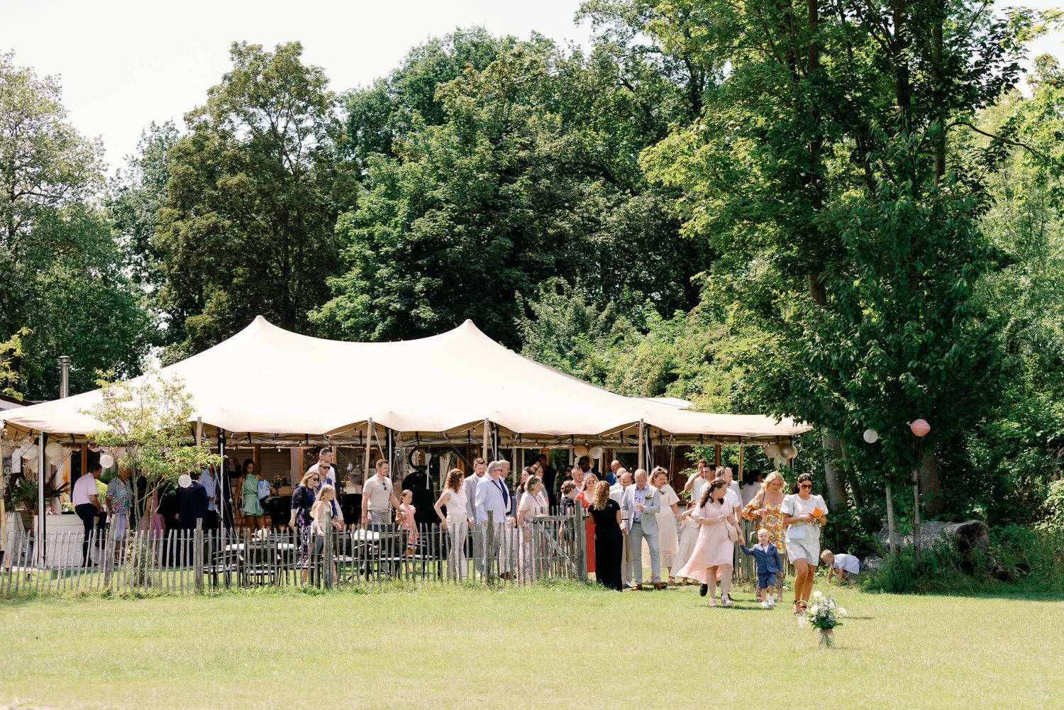 Panoramic view of an outdoor wedding reception setup with white tent canopies and guests gathered on a sunny lawn.