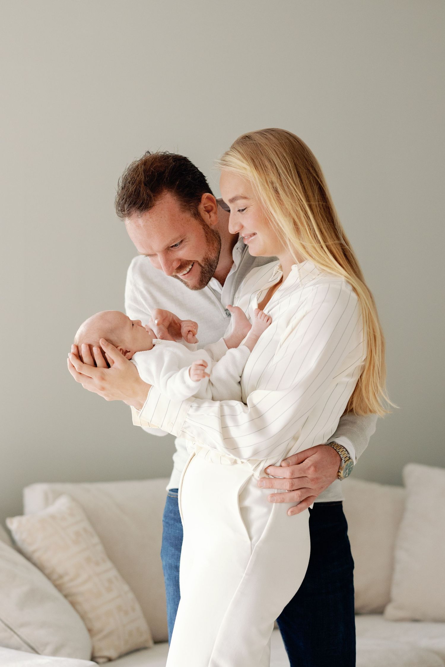 A couple in white clothing shares a tender moment while holding their newborn baby in a bright living room setting.