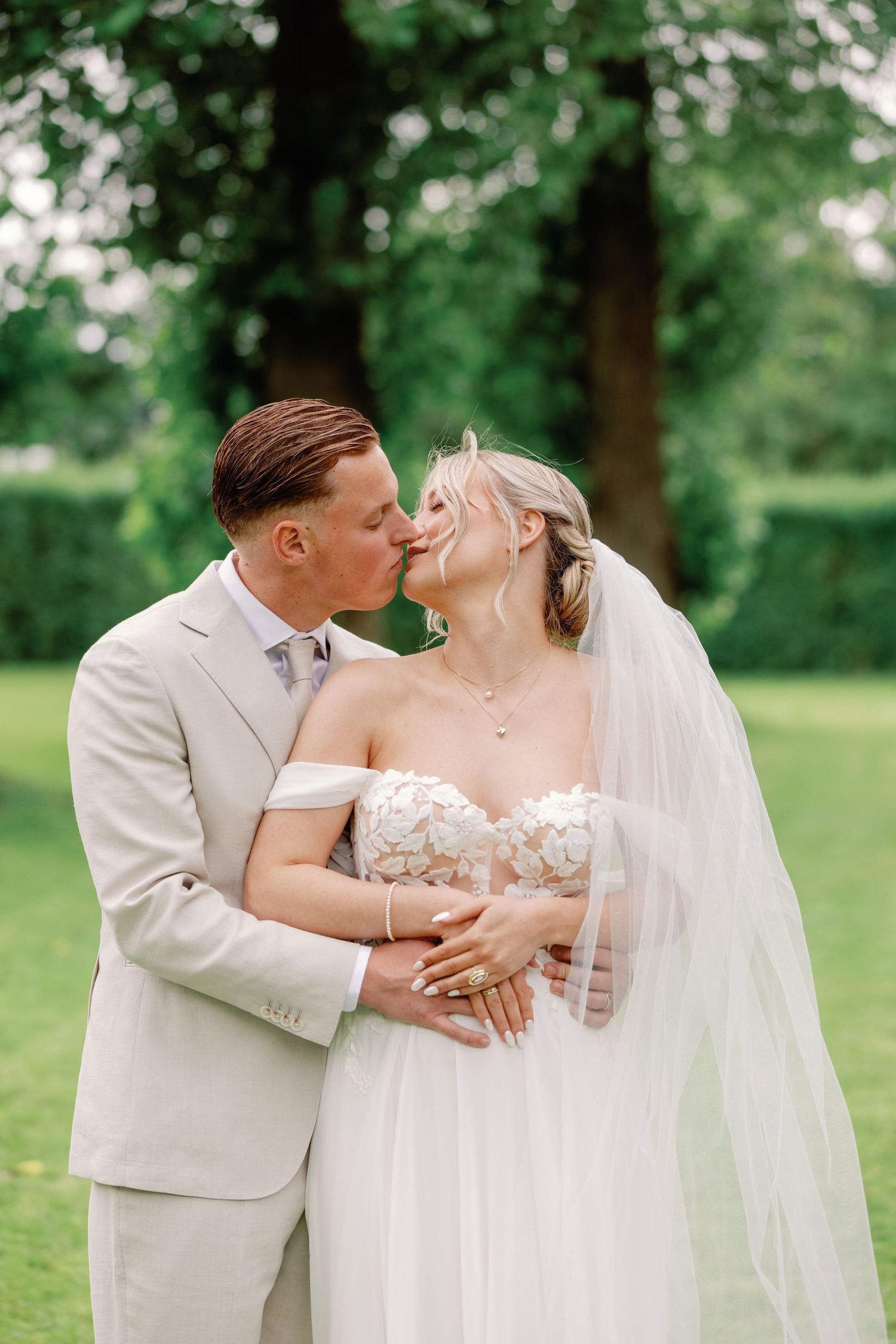 Romantic wedding couple embracing in a lush green garden setting wearing light colored attire.