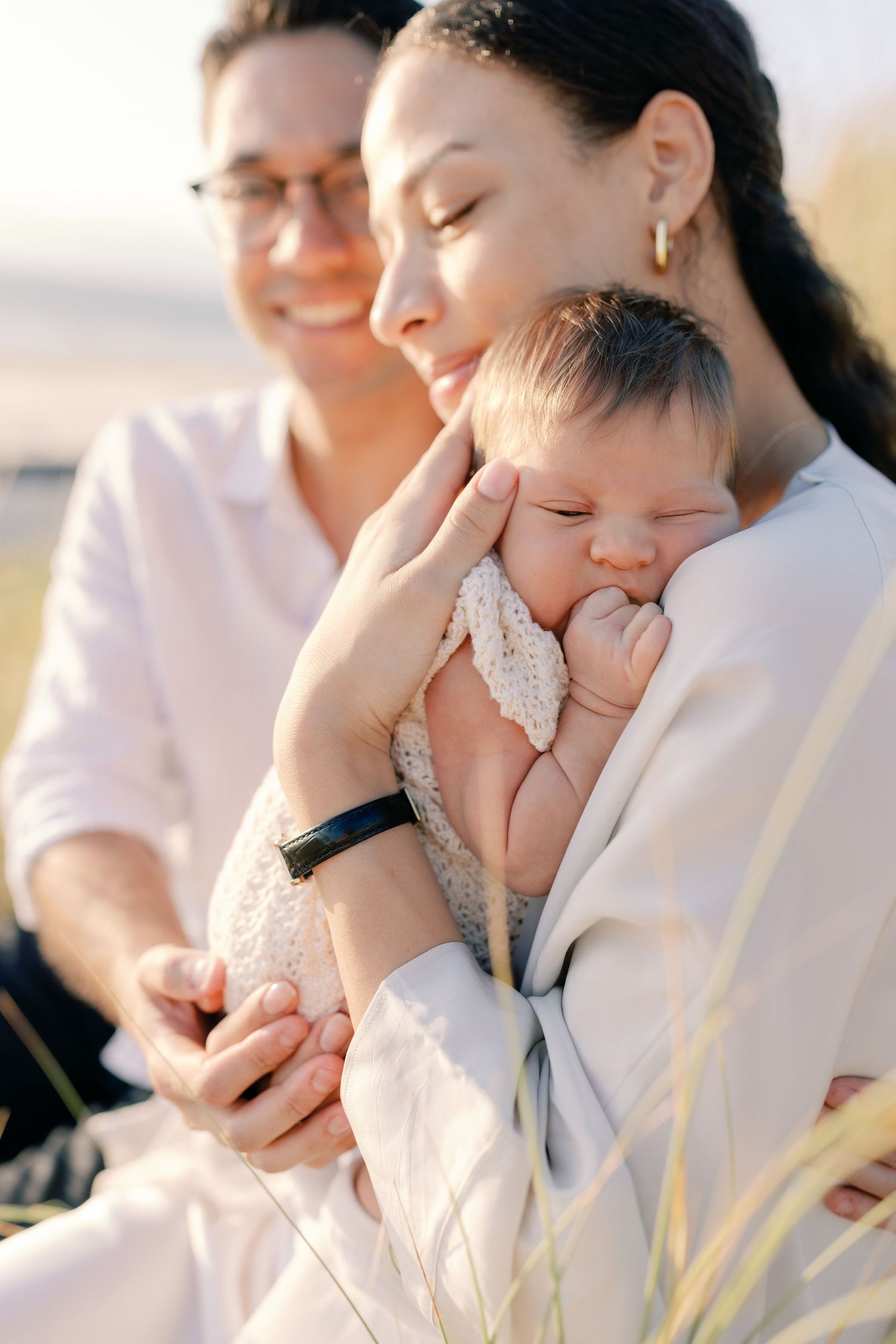 Parents in white clothing cuddle with sleeping infant against bright background.