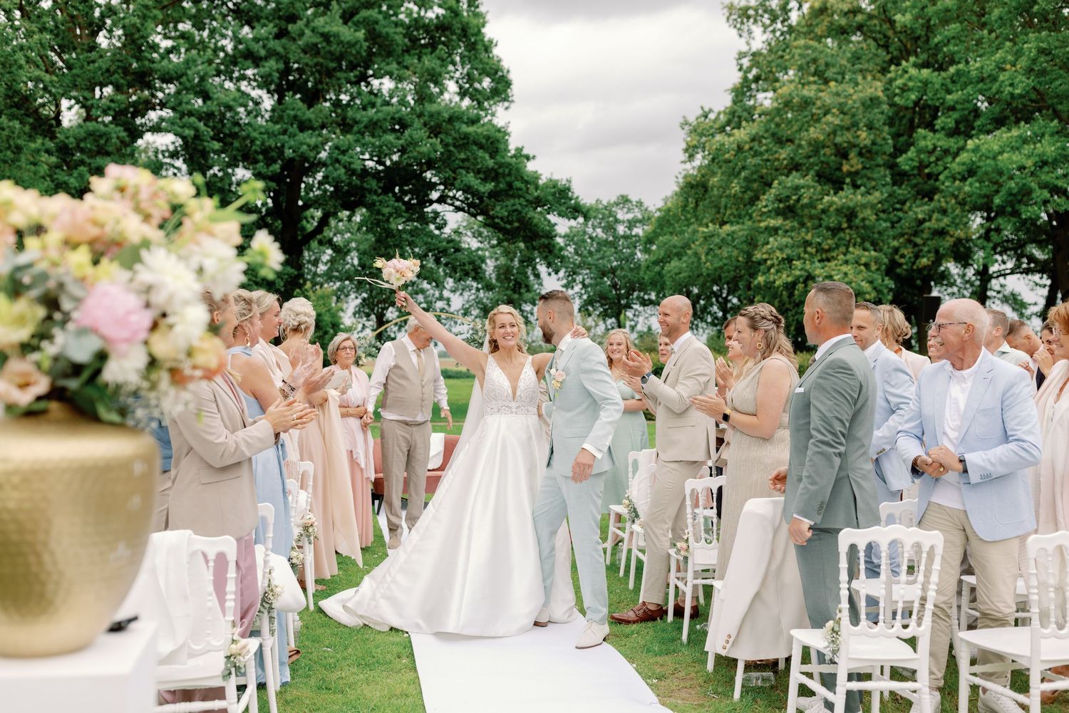Elegant outdoor wedding ceremony with white chairs and floral arrangements set against lush green garden backdrop.
