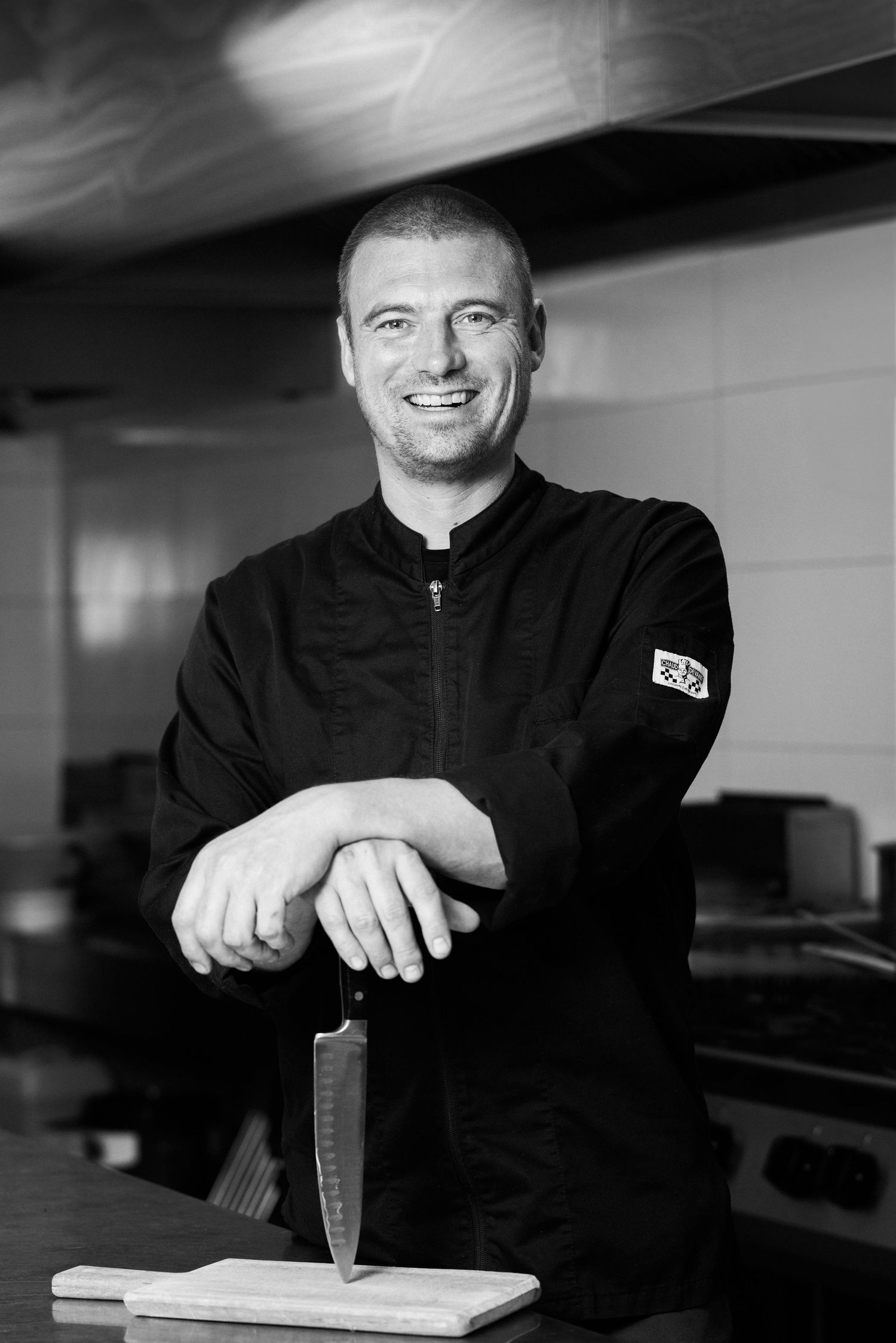 Black and white portrait of a chef in a dark uniform standing with a knife at a cutting board in a kitchen.