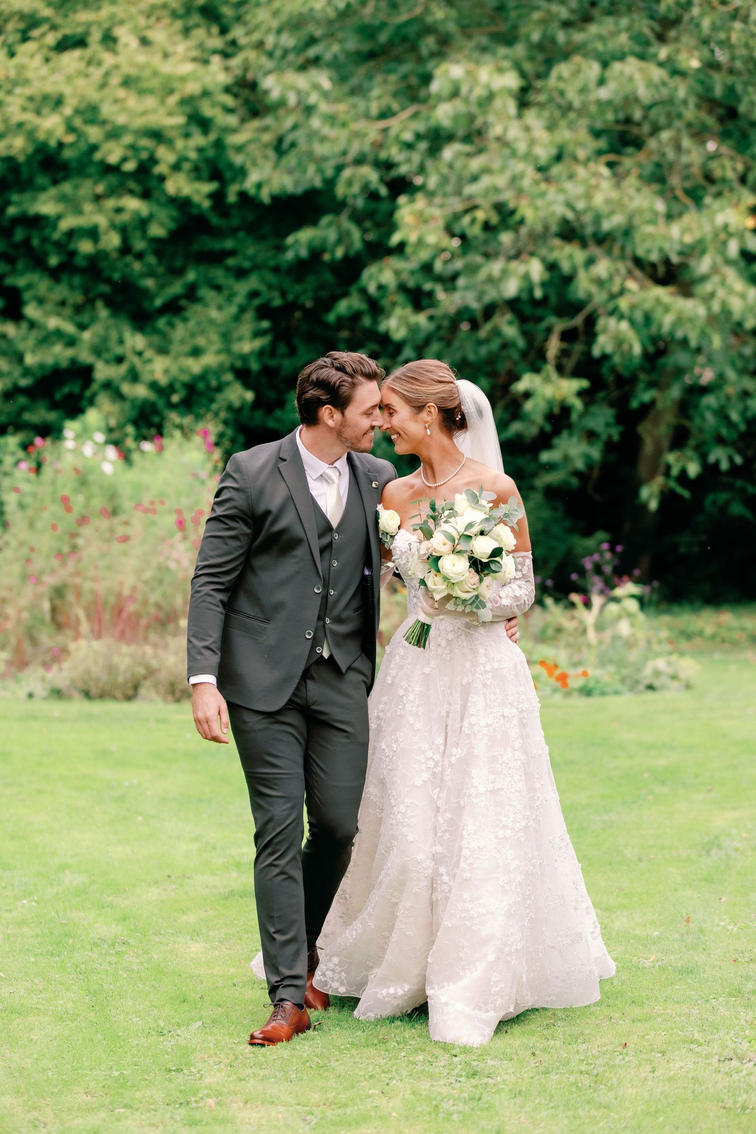 A romantic wedding couple walks together on a lush green lawn with white floral bouquet and classic formal attire.