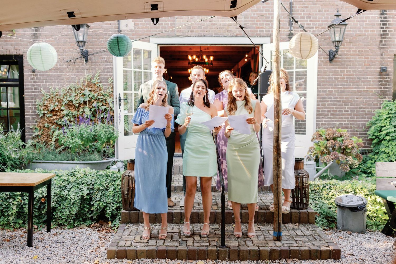 Four people celebrate at an outdoor garden party with paper lanterns and festive drinks on a brick patio.
