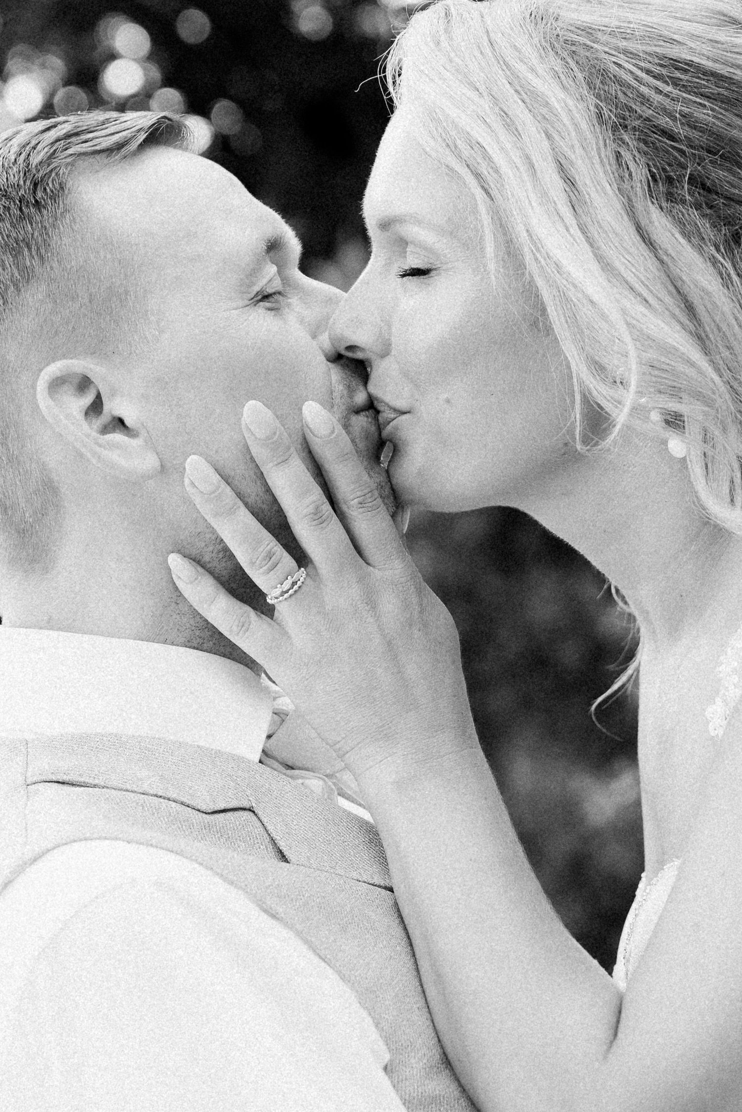 Black and white photo of a bride and groom sharing a romantic kiss, with her diamond ring visible on his cheek.