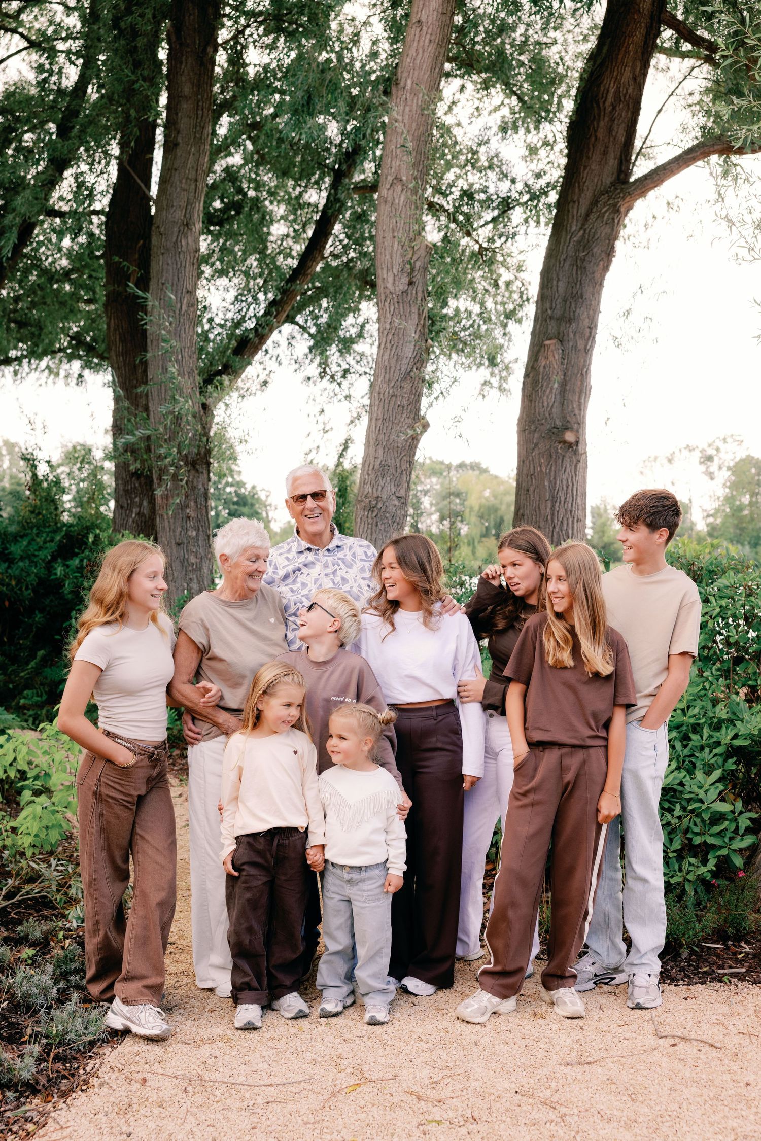 Multigenerational family of ten in neutral outfits laughing together on a gravel path beneath tall trees during an outdoor portrait session.