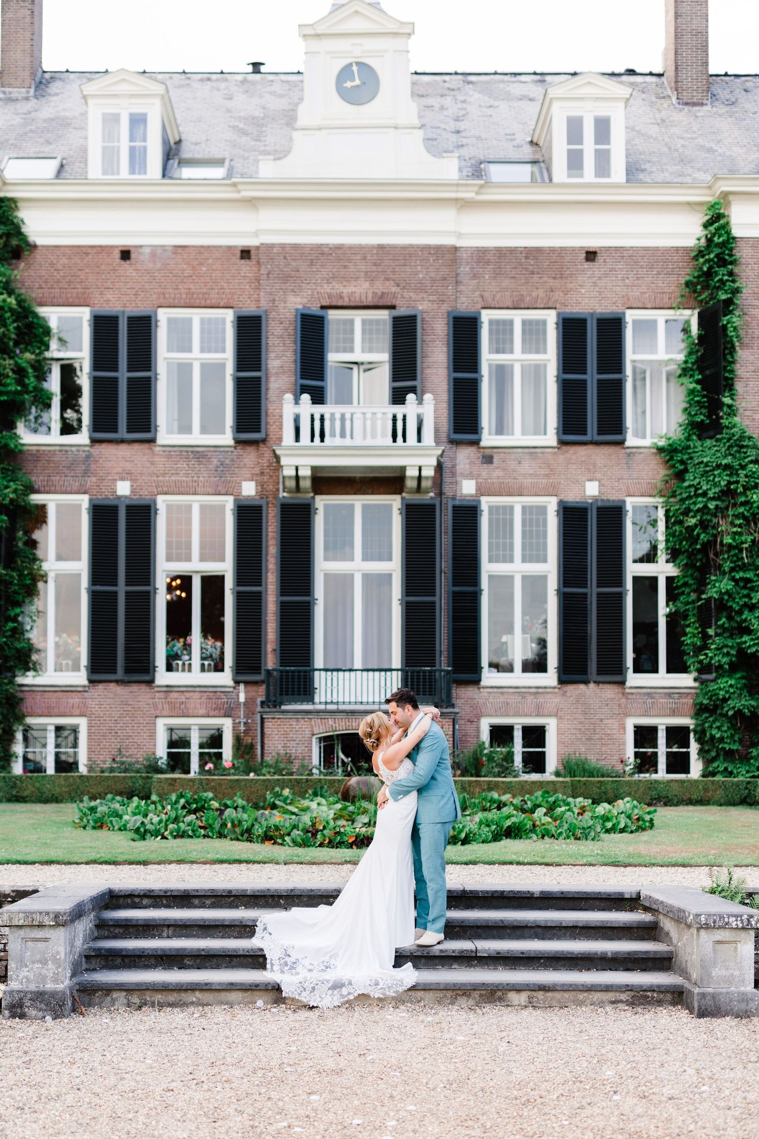 Elegant wedding couple embraces on stone steps in front of historic brick mansion with black shutters and ivy-covered walls.