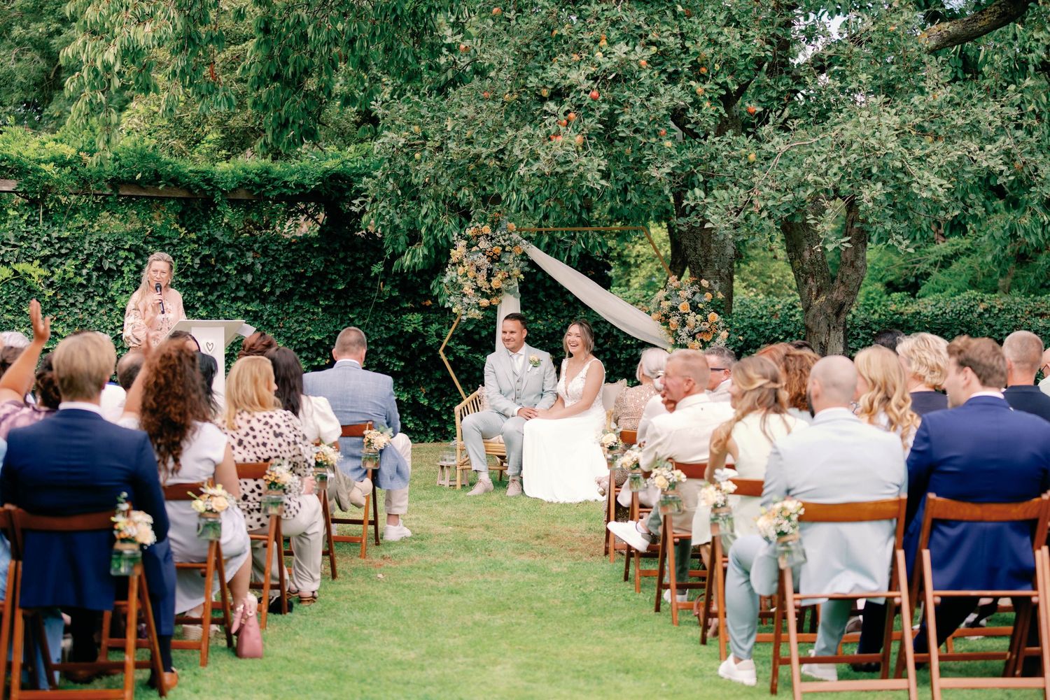 Panoramic view of an outdoor garden wedding ceremony with wooden chairs arranged in rows on a lush green lawn.