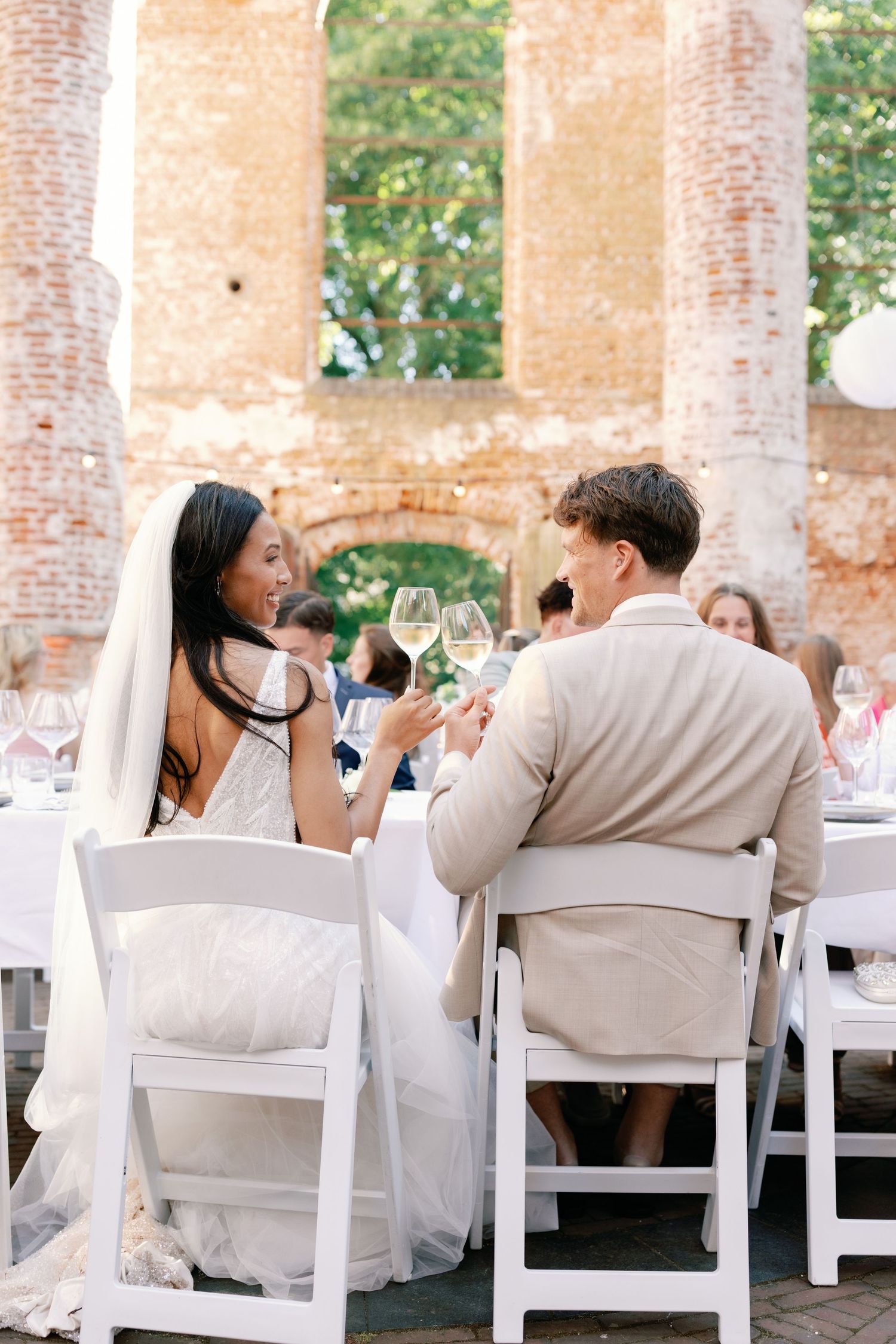 Wedding guests in white attire seated at elegant outdoor dining tables beneath rustic stone columns.