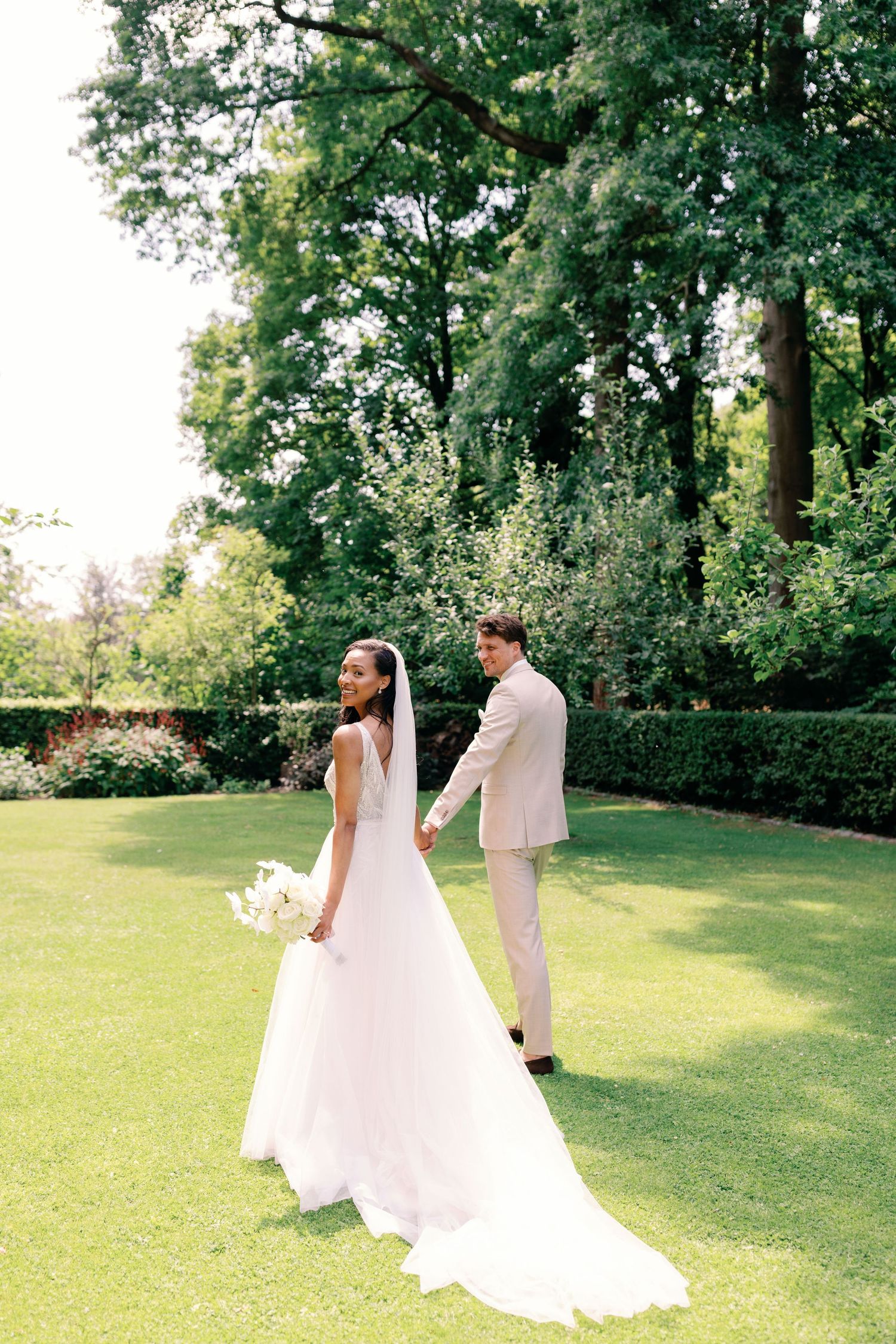 A couple in white wedding attire poses together on a lush green lawn surrounded by trees during their summer wedding.