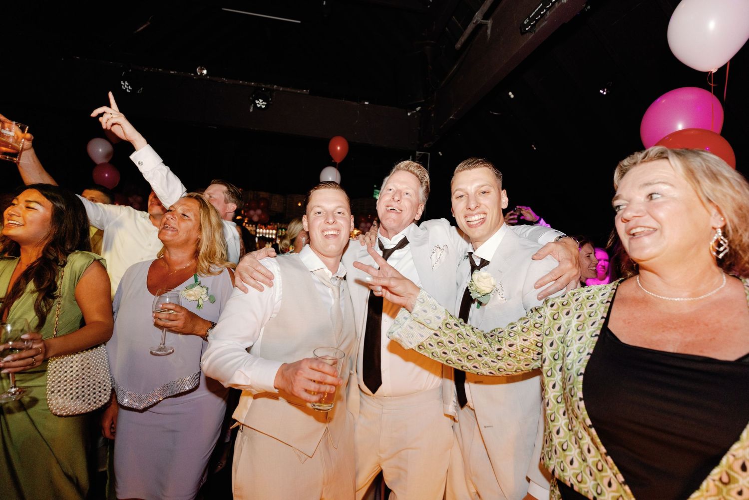 Party guests in light colored suits celebrate on a dance floor with balloons and festive atmosphere at a nighttime event.