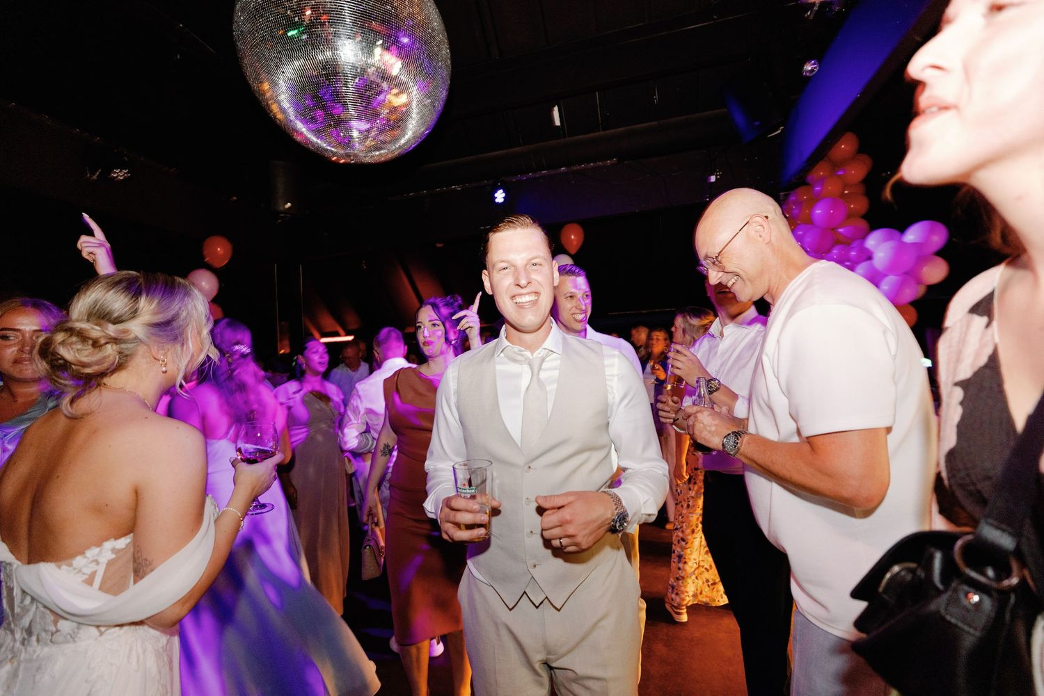 Party guests dance and celebrate under a disco ball with purple lighting at a nightclub event.