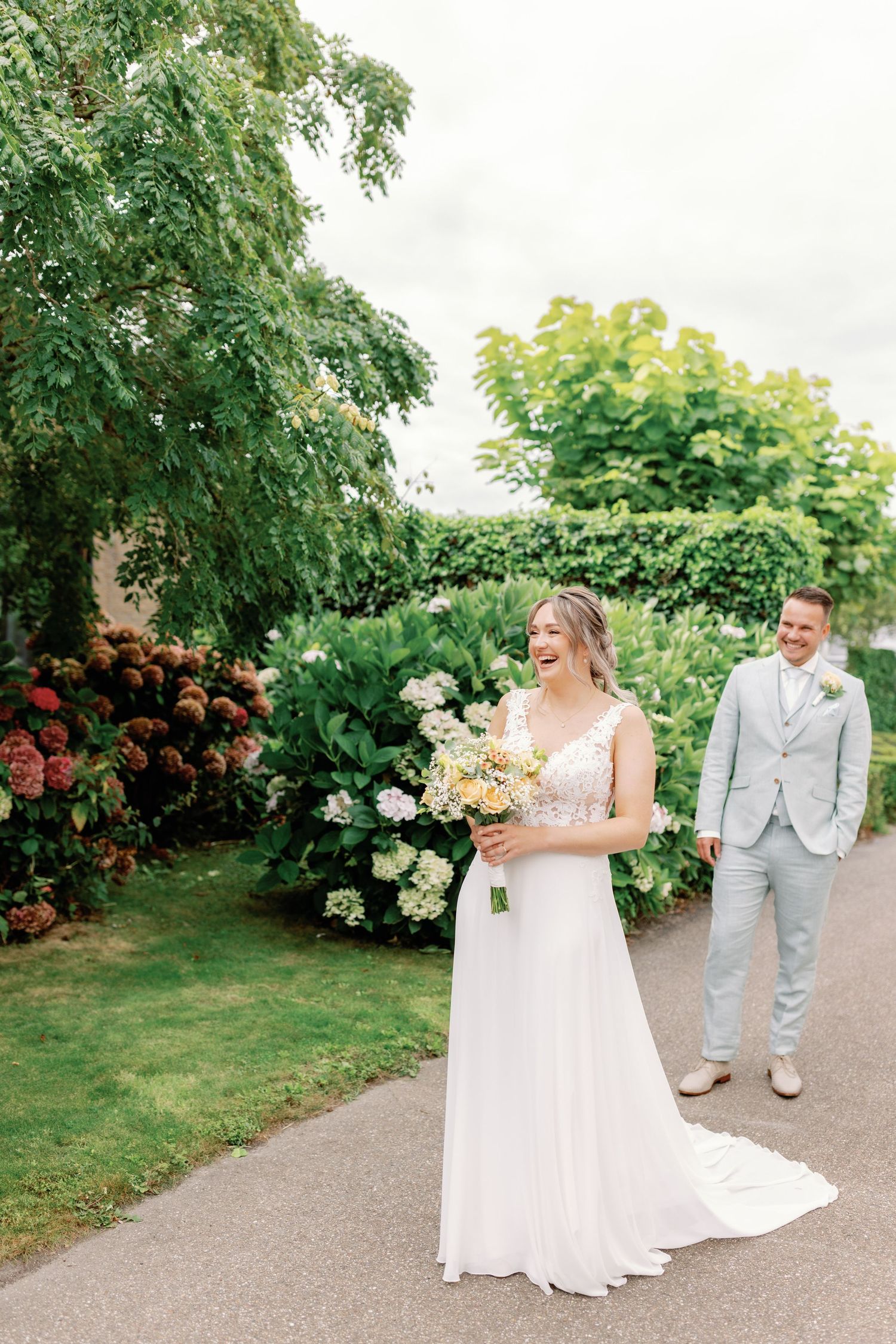 A joyful wedding couple shares a sweet moment on a garden pathway surrounded by summer blooms and trees.