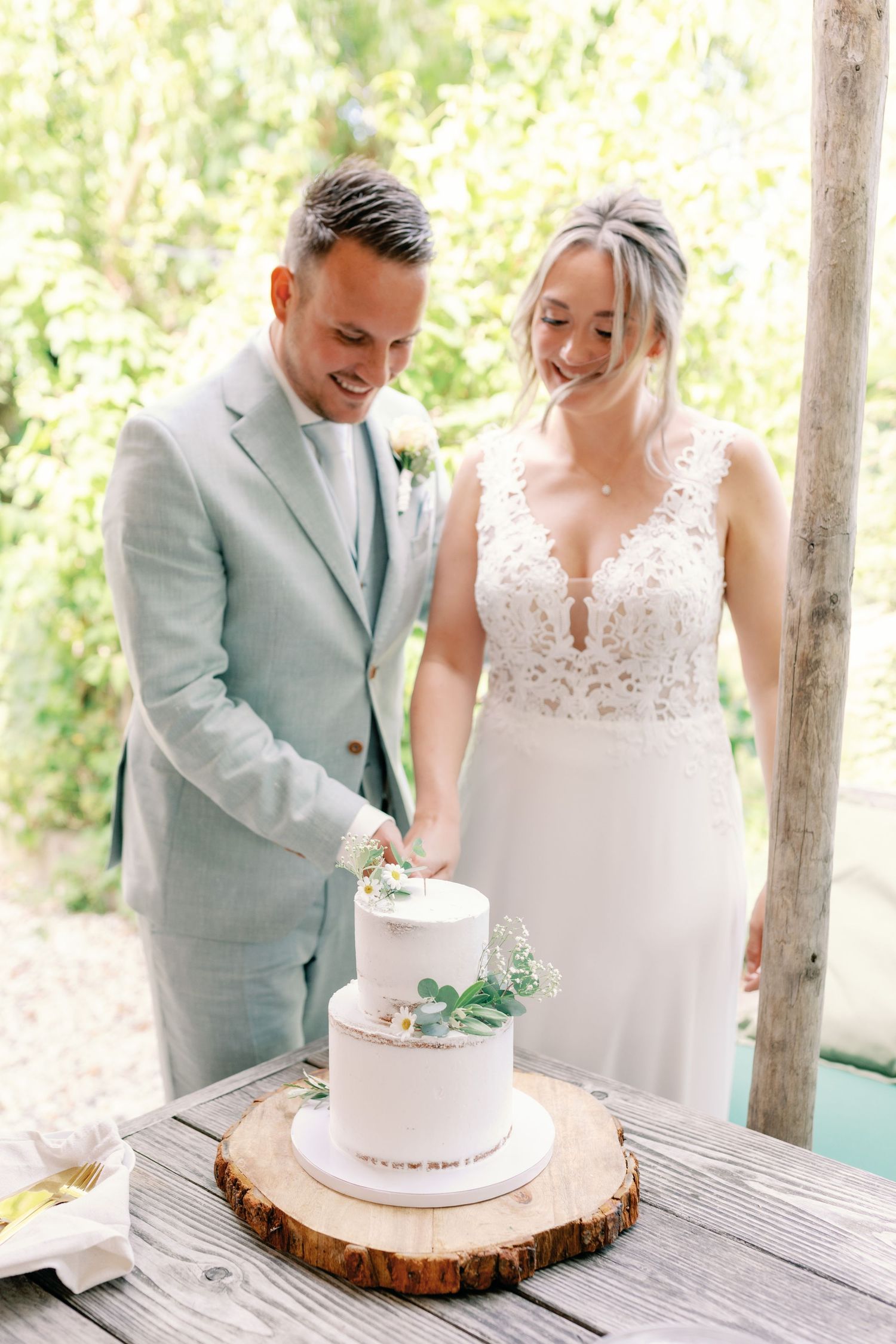 A couple in wedding attire stands together cutting their simple two-tier white cake decorated with greenery on a wood slice.