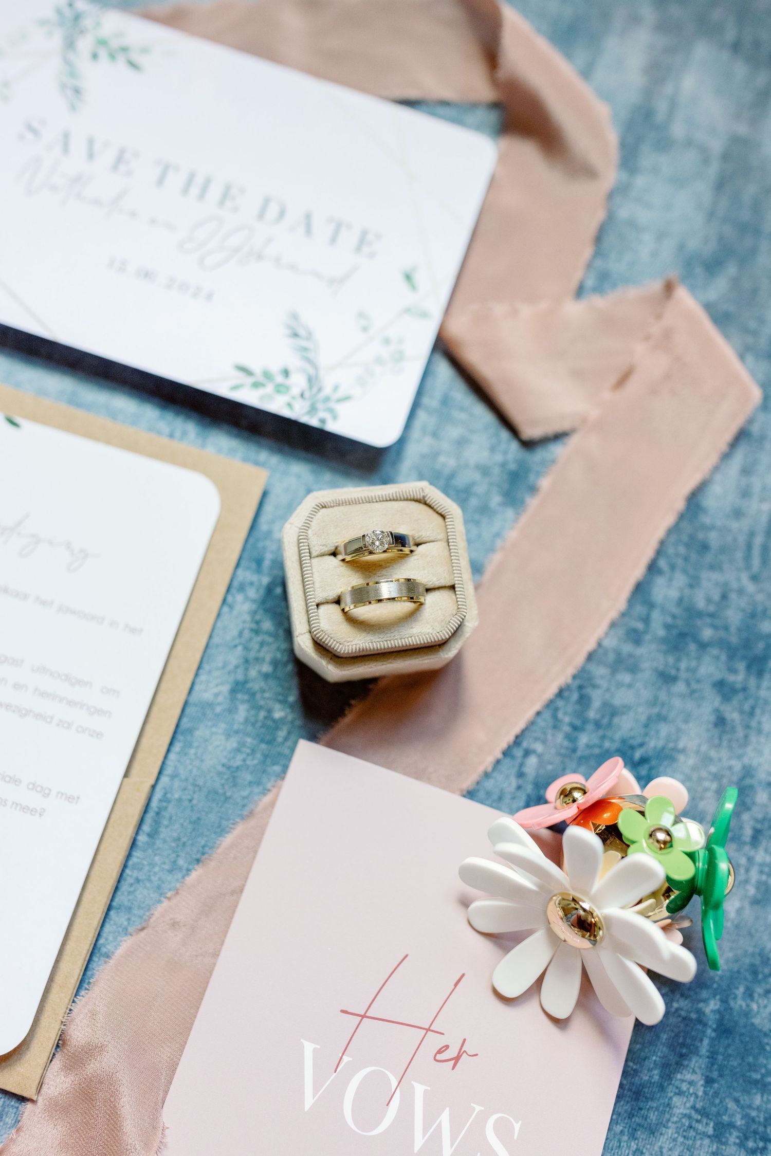 Wedding details including save the date card, velvet ring box with rings, and floral decor on blue textured background.