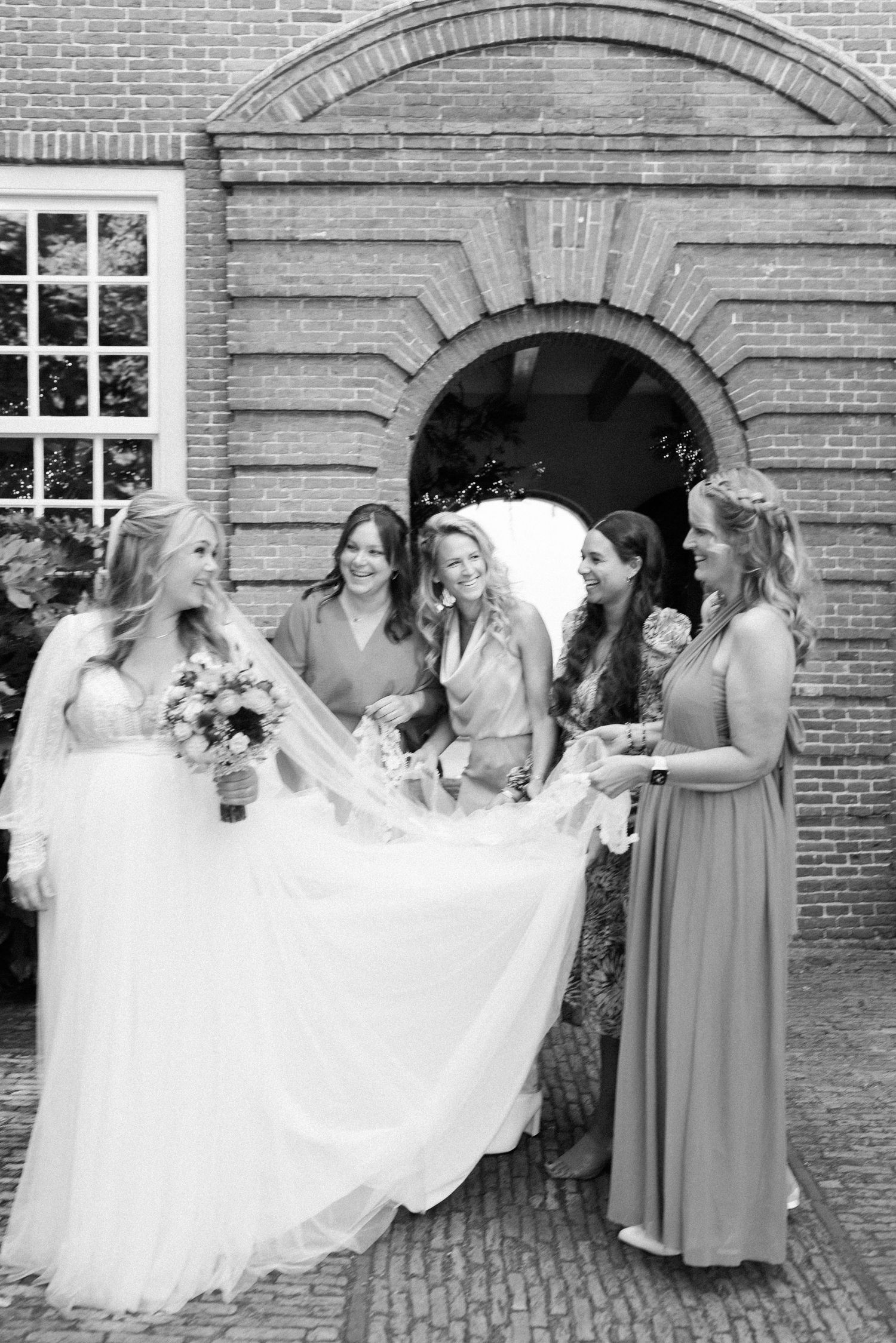 Bridesmaids in gray dresses help adjust a flowing white wedding gown veil against a brick building backdrop.