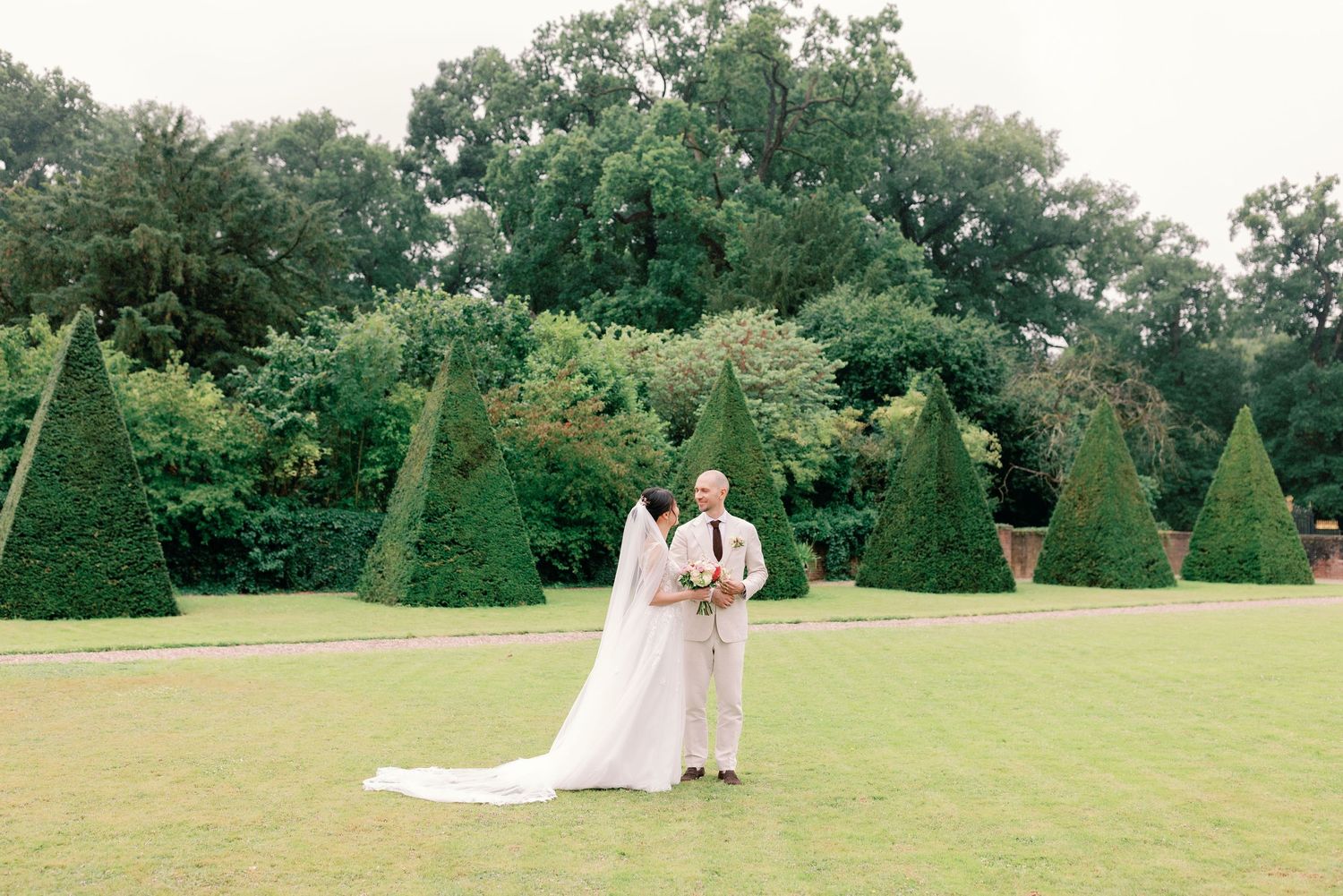 A couple embraces on a manicured lawn with sculpted topiary trees and lush greenery in the background.