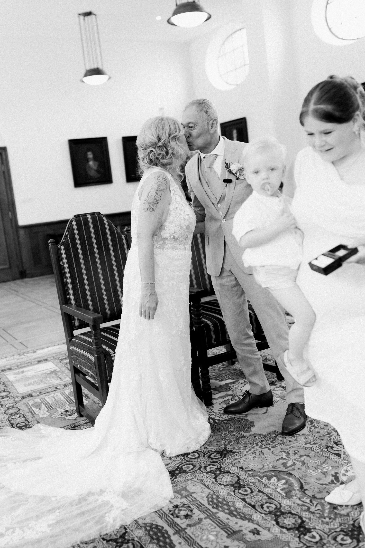 Black and white wedding photo of bride and groom kissing while woman holds baby in elegant venue.