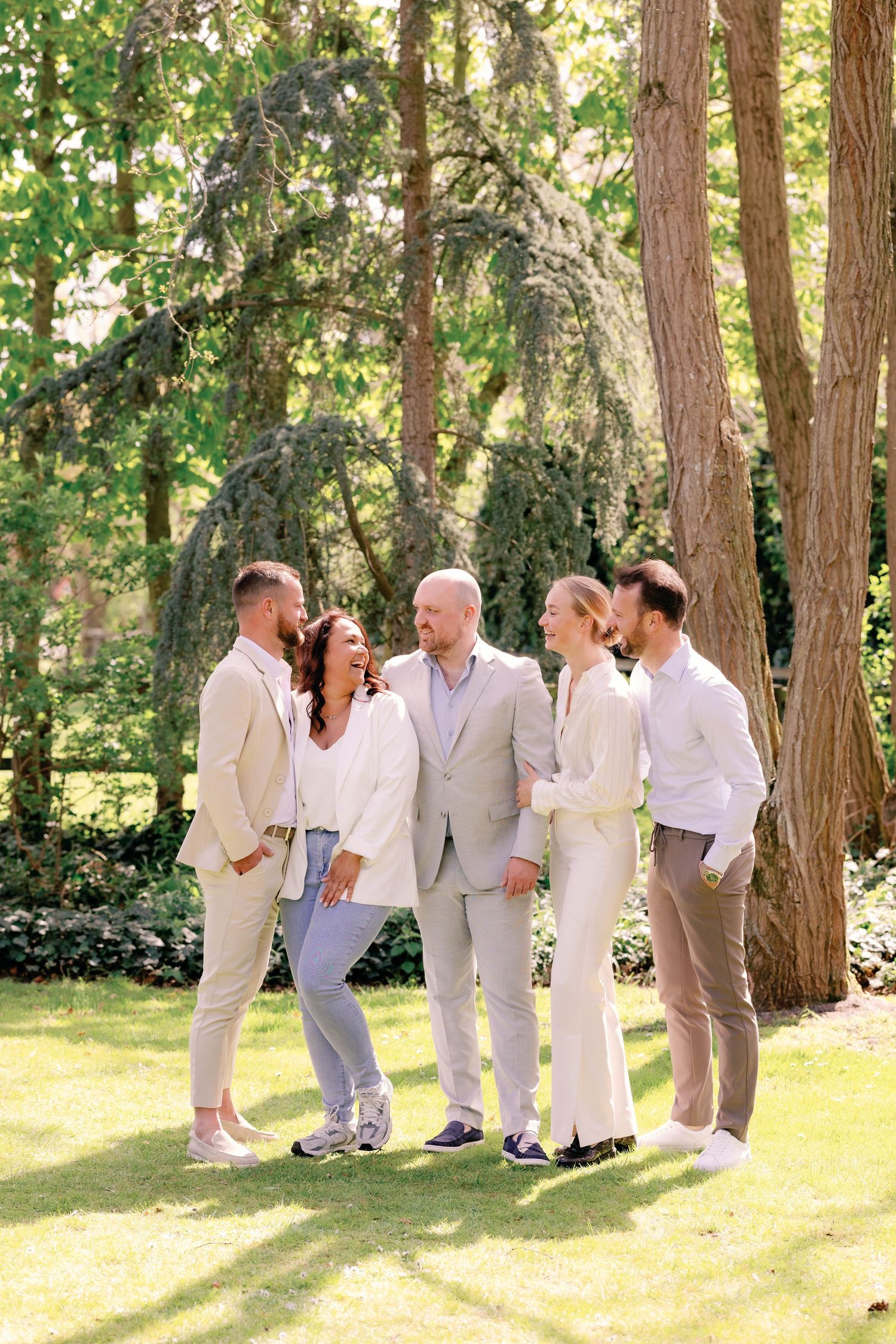 Five friends in neutral outfits laughing together on a sunny green lawn surrounded by tall trees.