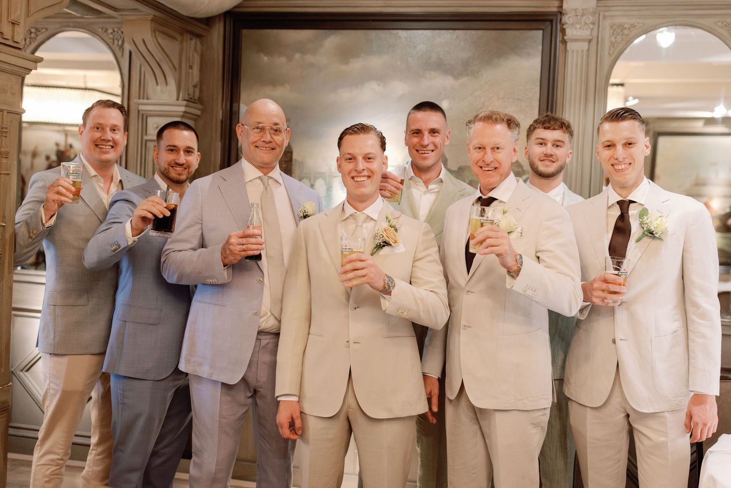 Groomsmen in matching tan suits pose together in front of decorative wall panels at a formal event.