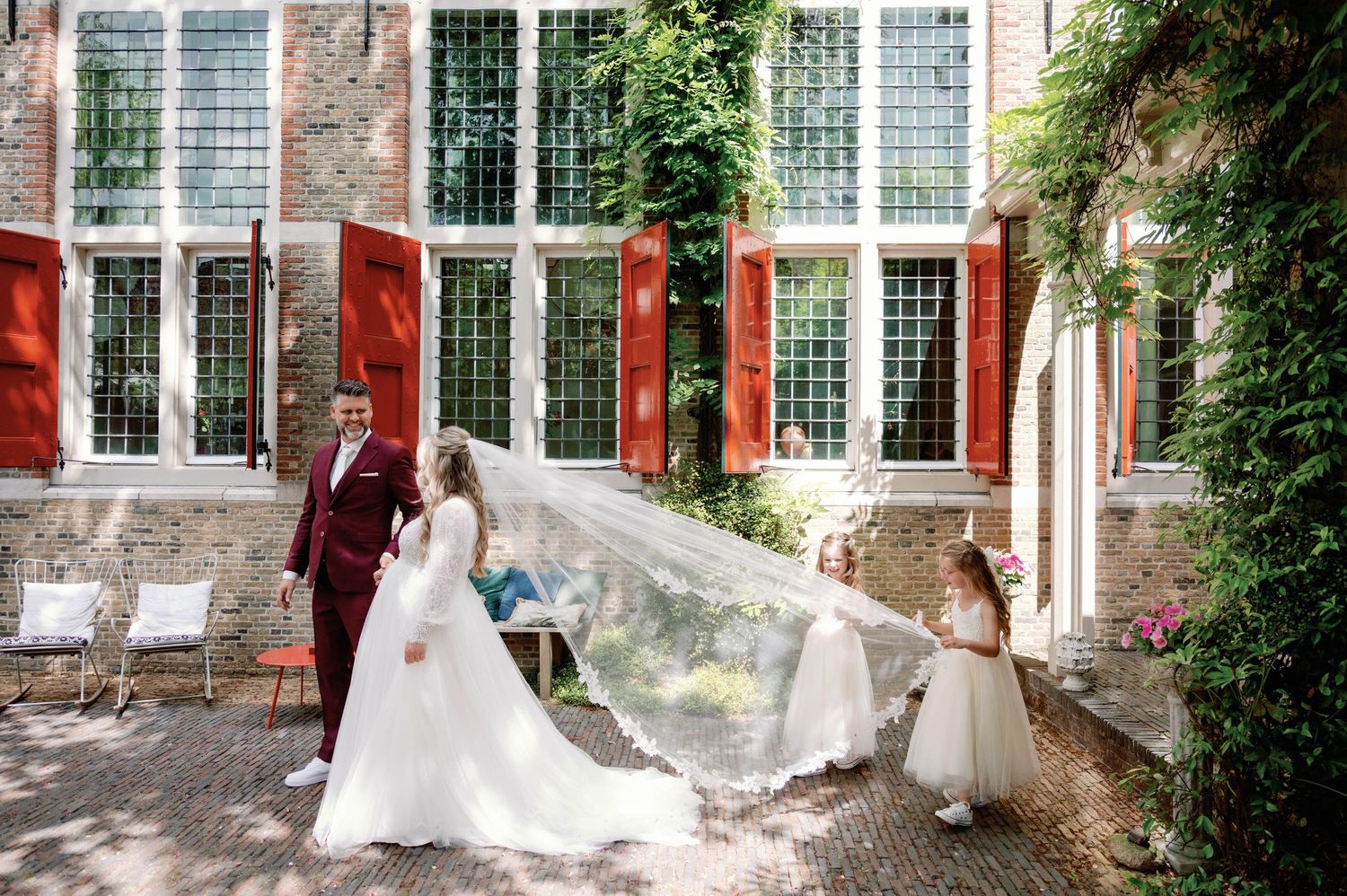 A wedding party poses in front of a historic stone building with red trim and large windows while a veil flows in the breeze.