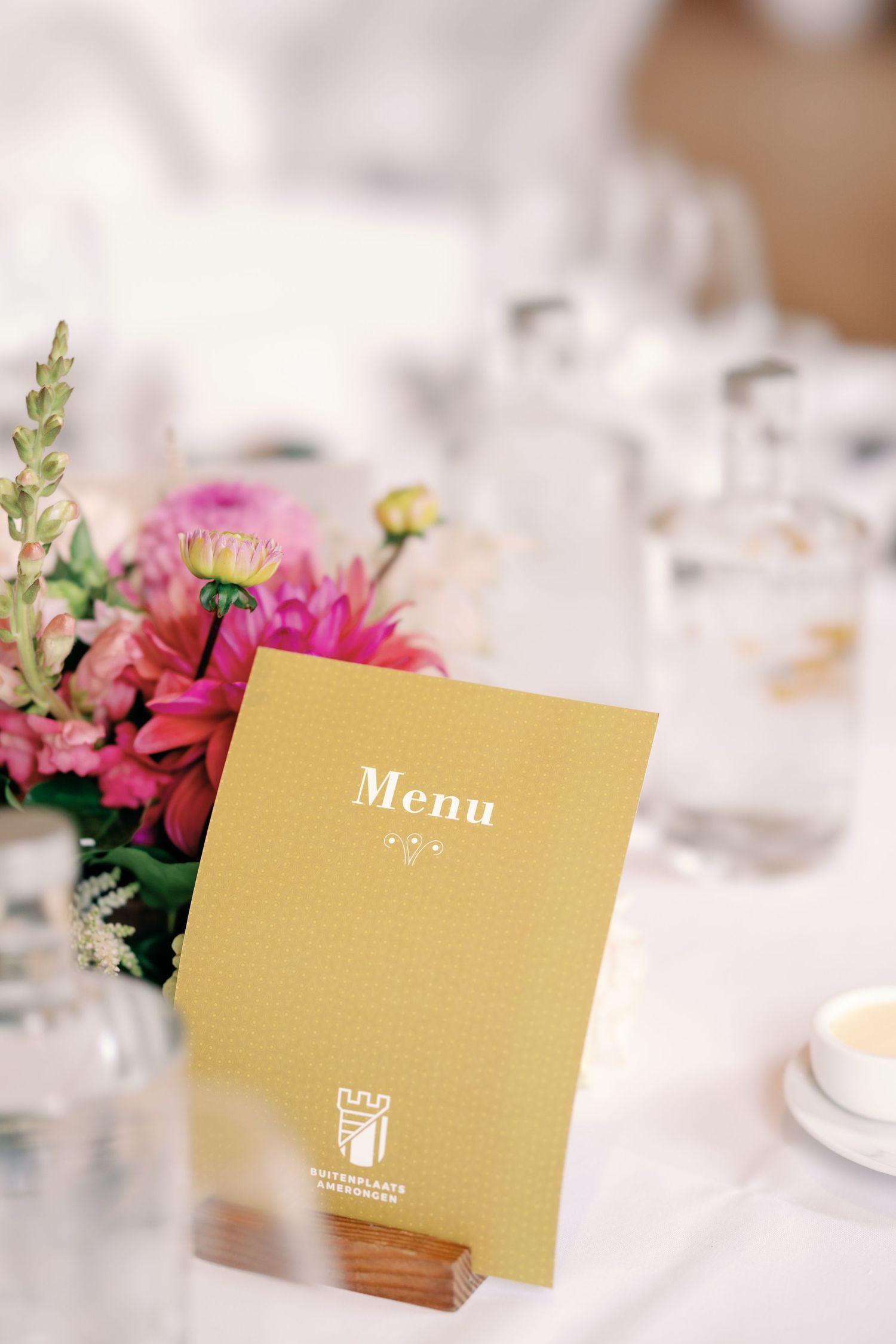 A gold menu card stands on a table with pink floral centerpiece and glassware at an elegant event setting.