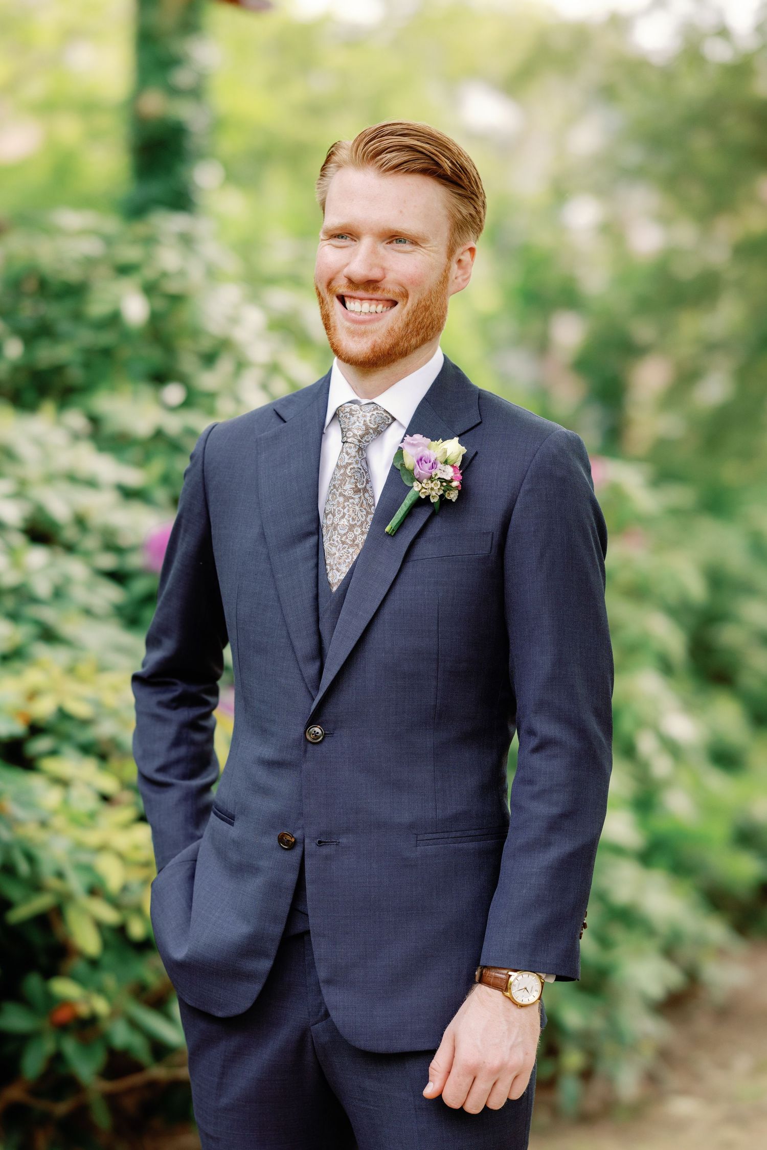 A groom in a navy suit and patterned tie smiles joyfully during outdoor wedding portraits in a garden setting.